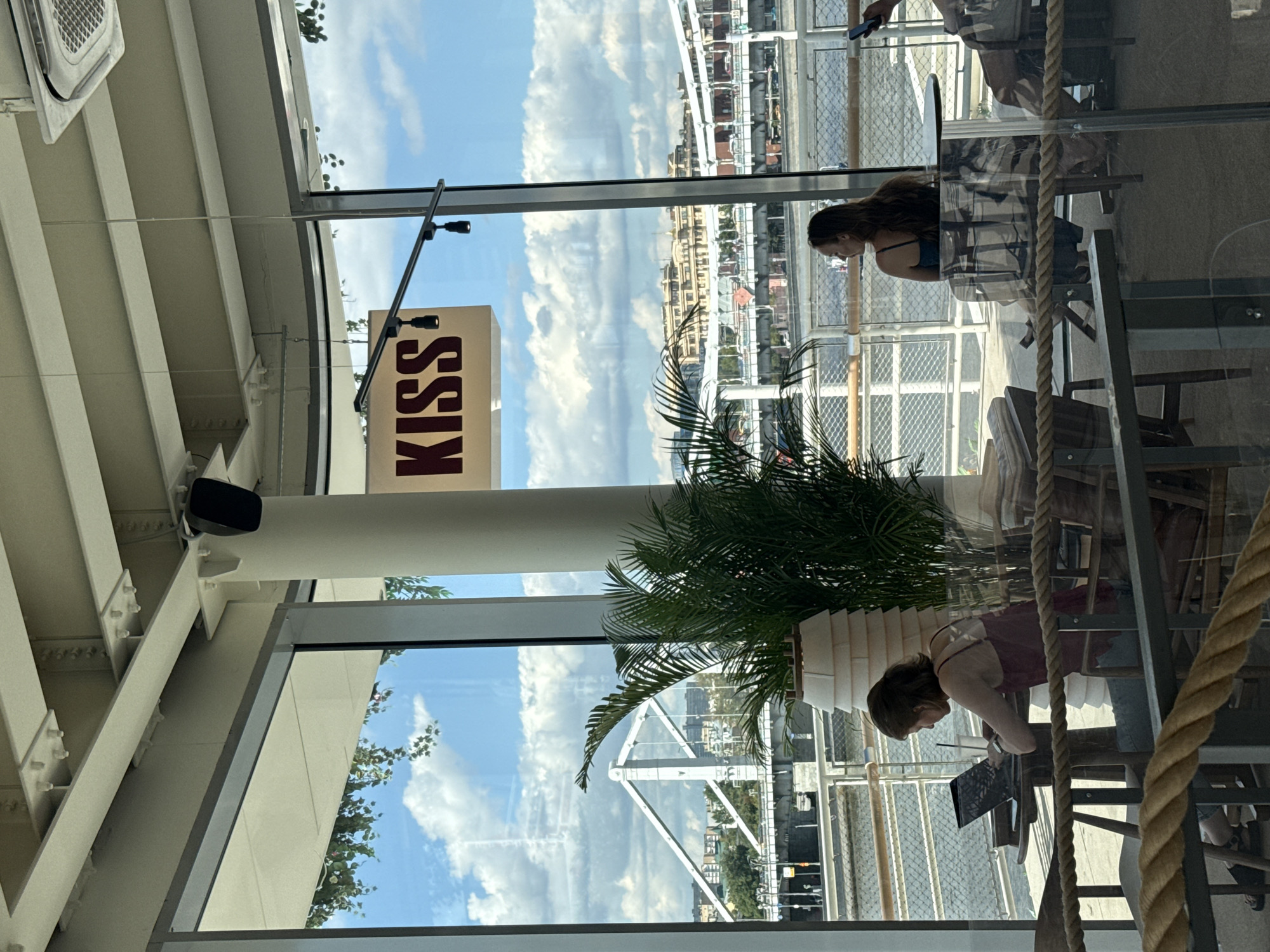 The image depicts the interior of a modern cafe with large windows offering a scenic view of the Moskva River and the Crimean Bridge in Moscow, Russia. A woman with long brown hair is seated at a table on the left, working on a laptop. She is wearing a dark pink tank top and jeans. A large potted palm is positioned between her and the window.
In the background, across the river, a cityscape with buildings and a bridge is visible under a partly cloudy sky. A sign with the word "KISS" in red lettering against a cream background is mounted on a white pillar in the mid-ground. To the right, another woman with long brown hair is seated at a circular table, looking towards the window. She is wearing a dark blue dress. Reflections on the glass indicate that this is likely a daylight scene. The cafe appears to be furnished with wooden tables and chairs. A thick rope barrier runs across the foreground.
