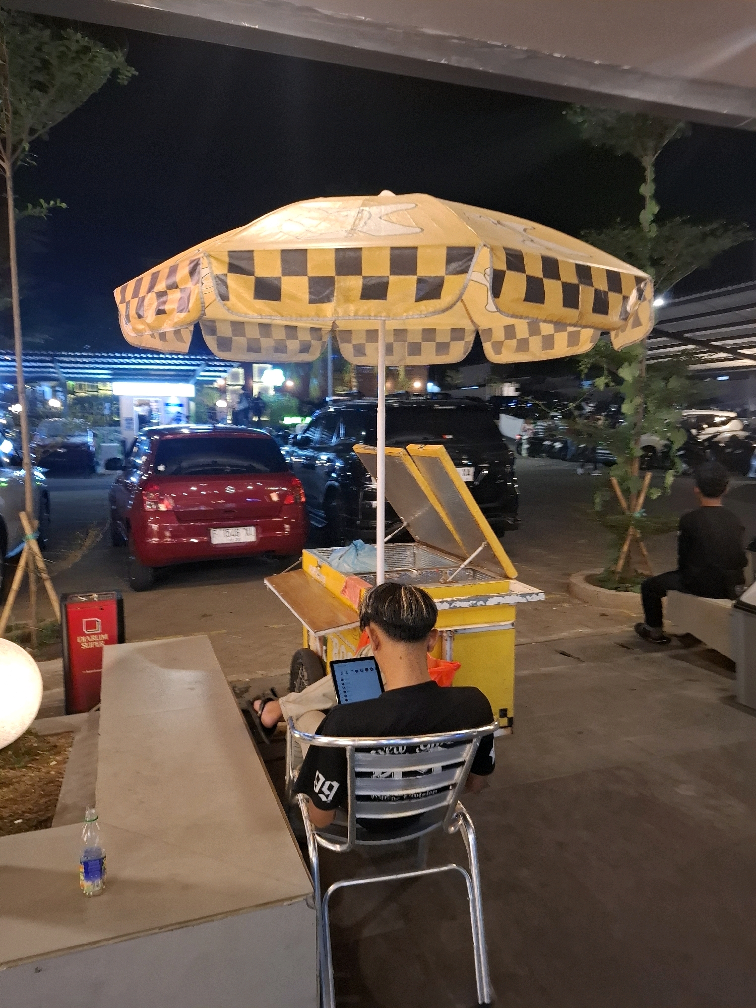 The image shows an outdoor seating area at night, likely at a food stall or market in Cianjur, Indonesia. A person is seated in a metal chair, facing away from the camera and looking at a tablet. They are wearing a black t-shirt with white lettering. In front of the person is a yellow food cart with a checkered yellow and black umbrella. A red trash can is visible to the left of the cart. In the background, several cars are parked, including a red car with a license plate that appears to read "F 1545 XL". The environment is lit by artificial lights, and there are trees and an awning overhead.