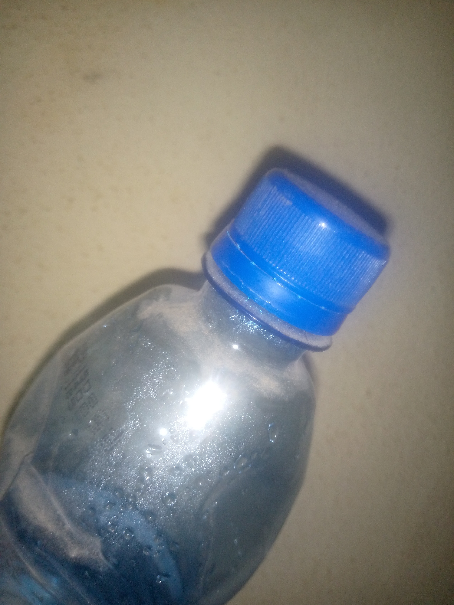 A close-up, angled shot shows the top portion of a clear plastic water bottle with a blue screw-on cap. The bottle appears to have condensation on its surface, indicating it may be cold. There is some visible dust on the bottle and the cap. The background is a neutral, textured surface, possibly a table or wall, that is out of focus. Faint markings or text are visible on the bottle, but they are illegible.
