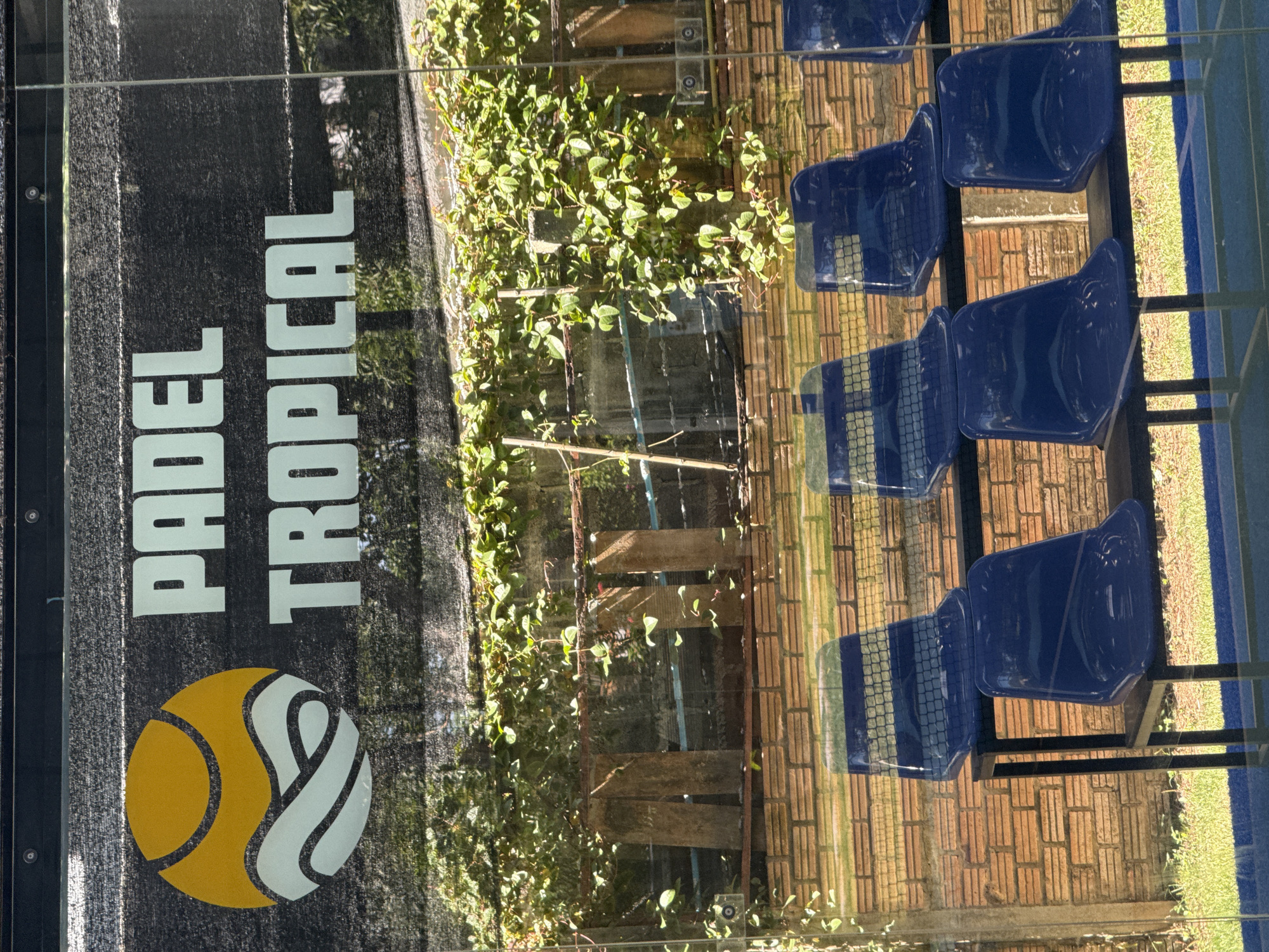 The image shows a close-up view of a glass barrier with reflections of blue plastic stadium seats. Behind the seats is a brick wall overgrown with green vines. Above the seats and wall, a black mesh material is visible with a logo that appears to be for "PADEL TROPICAL". The logo consists of a yellow circle with white wave-like shapes inside. The lighting suggests it is daytime, with sunlight casting shadows and reflections. The location is identified as Ban Bang Rak, Thailand. There are no people visible in the frame.