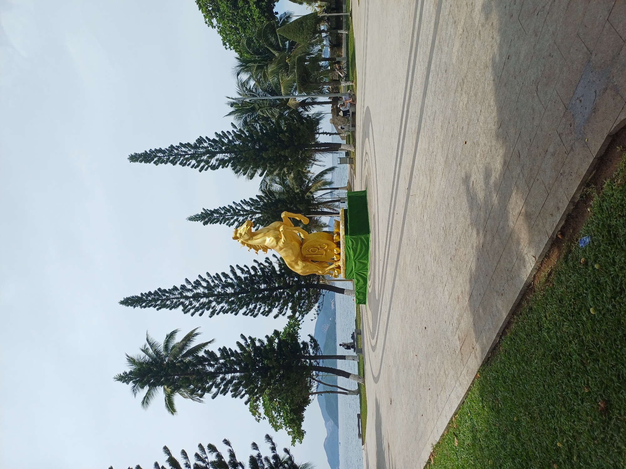 This image depicts a golden statue of a horse rearing on its hind legs, positioned on a green base. The statue is located in a paved outdoor area, possibly a park or promenade, with a grassy area on the left and trees and the sea in the background. The overall scene suggests a public space designed for leisure and appreciation of art, possibly in a coastal city. The statue is centrally placed, drawing the viewer's eye, and the surrounding trees and water create a serene and natural backdrop. The paving stones in the foreground are laid in a patterned arrangement, suggesting a well-maintained urban environment. The statue appears to be the main focal point, with a few individuals visible in the background, suggesting a relatively calm and uncrowded setting. The presence of palm trees and the proximity to the sea indicate a tropical or subtropical climate, consistent with Nha Trang, Vietnam. The golden hue of the horse statue, combined with the lush greenery and the expansive seascape, creates a visually appealing and inviting atmosphere.