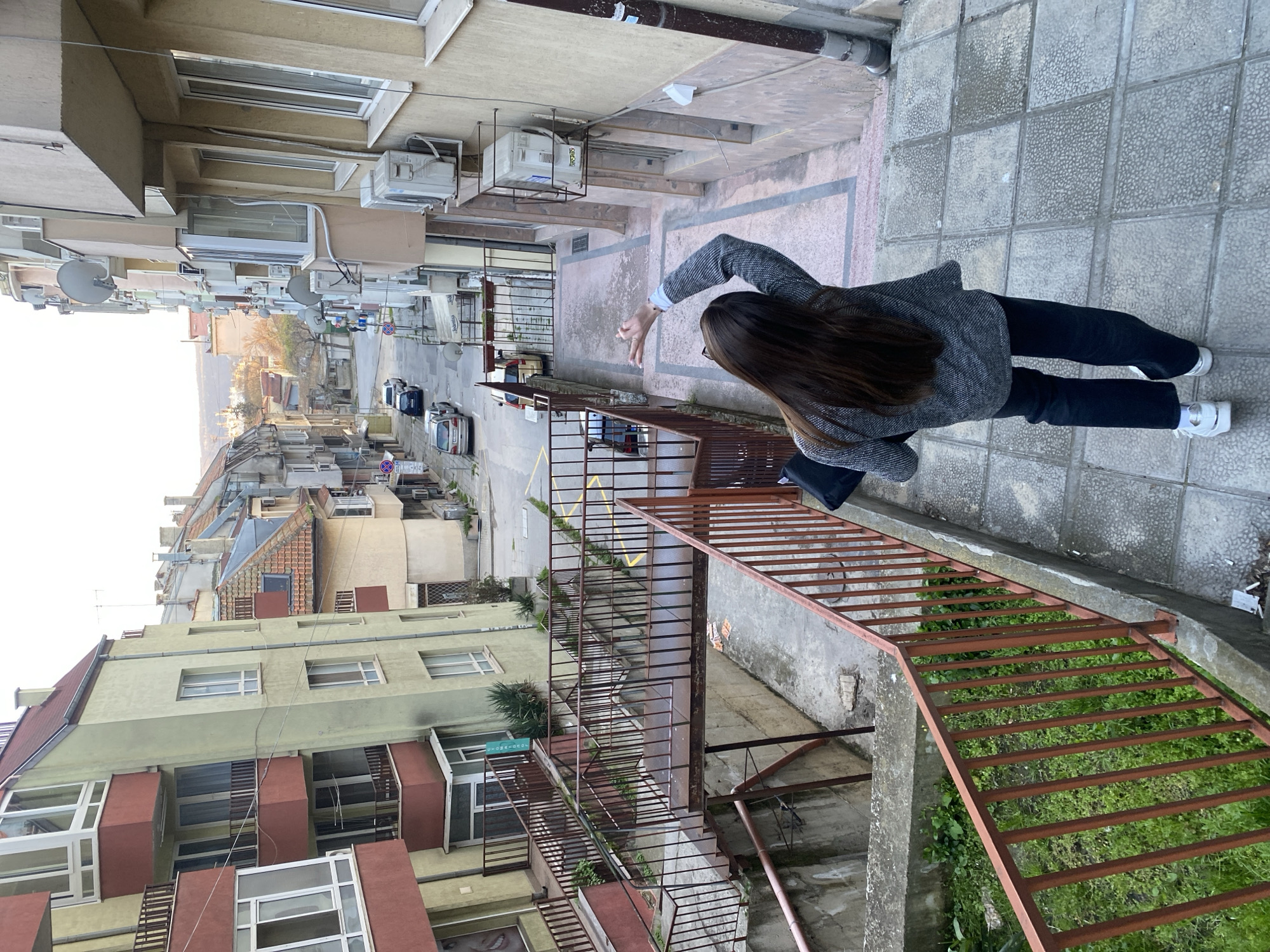 A woman with long dark hair is standing on a paved area in Varna, Bulgaria, leaning over a railing. She is wearing a gray jacket, black pants, and white sneakers. She is facing the street, and her right hand is extended towards it. The street has cars parked along it. Buildings line both sides of the street. Below the railing where the woman is standing is a set of metal stairs leading down to a lower area with vegetation. A satellite dish is visible on a building across the street. Air conditioning units are mounted on the building adjacent to the area where the woman is standing. The image is shot from an elevated perspective.