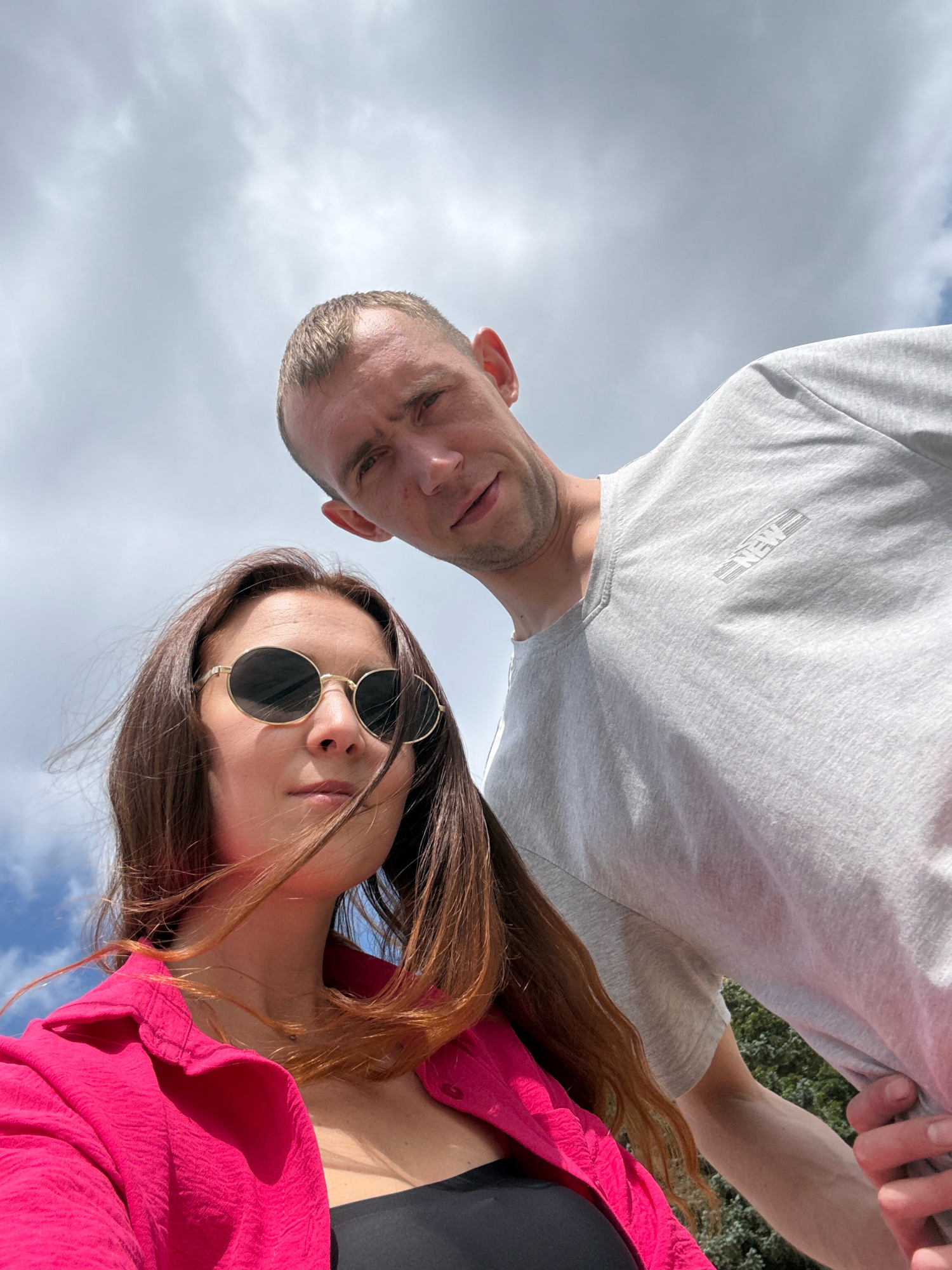 In a low-angle selfie against a cloudy sky, a couple is captured outdoors. A man with short hair and a light gray t-shirt is positioned behind and slightly to the right of a woman. The woman, with long brown hair and wearing round, gold-rimmed sunglasses and a vibrant pink shirt over a black top, is in the foreground, looking towards the camera. The background consists of the sky and some greenery. The overall impression is a casual, perhaps spontaneous, moment captured by the couple themselves, possibly in a park or outdoor recreational area. No specific landmarks in Maykop, Russia, are discernible from the image.