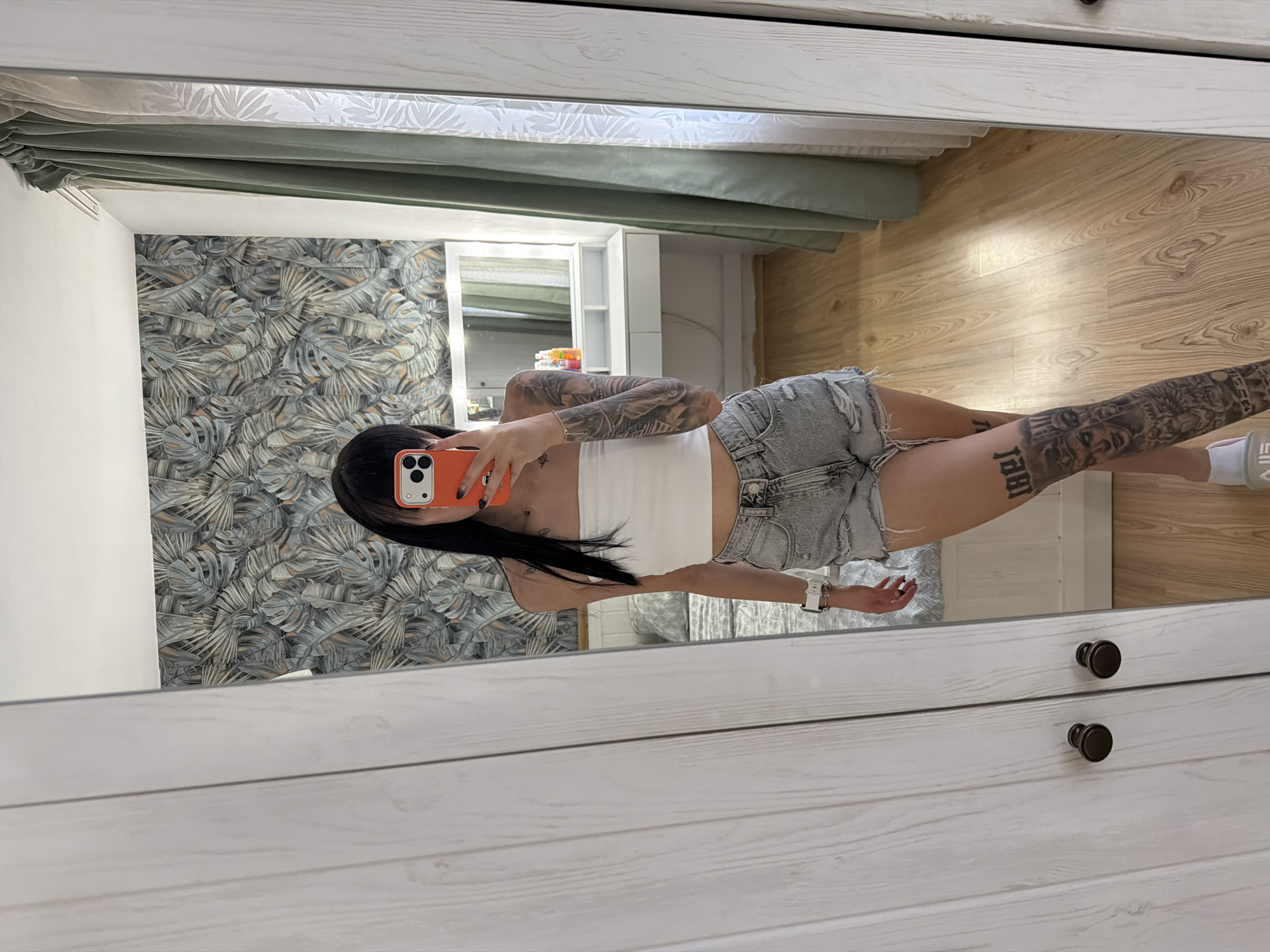 A woman with long dark hair is taking a mirror selfie in what appears to be a bedroom. She is wearing a white tube top and distressed denim shorts. Her left arm and right leg are heavily tattooed. She is holding an orange smartphone. In the reflection, a vanity with a lighted mirror and various bottles can be seen. Behind the vanity, there is a wall with a tropical leaf pattern wallpaper. To the right of the vanity, there are green curtains. The floor is made of light-colored wood. The woman is standing in front of a large, white wardrobe with two doors. The image does not provide any information about the location in the city of Plovdiv, Bulgaria.