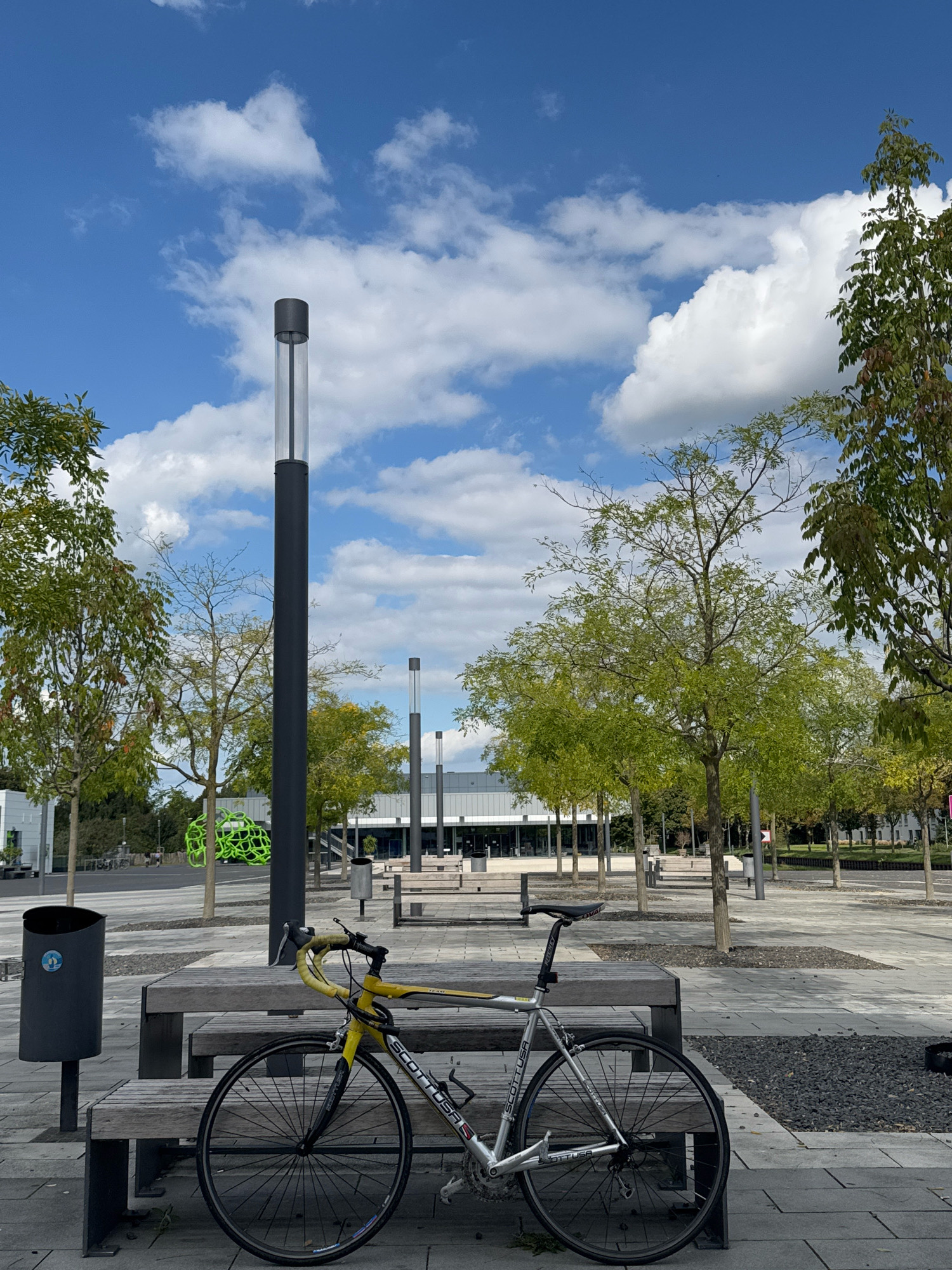 This image captures an outdoor scene in the city of Cleves, Germany. In the foreground, a silver and yellow road bicycle is parked next to a wooden bench. The bicycle has a yellow handlebar tape and "SCOTT USA" branding visible on the frame. To the left of the bench, a dark grey trash can with a blue circular logo is present. The bench and trash can are situated on a paved area with square sections of dark gravel.
Behind the bench, several trees with light green foliage are planted in rows, creating a somewhat urban park-like atmosphere. In the background, a modern building with a white facade and dark windows is visible. A tall, dark grey lamppost with a cylindrical light fixture stands prominently in the middle ground. Further back, several more lampposts are visible, receding into the distance. The sky above is blue with scattered white cumulus clouds. The overall environment suggests a public space, possibly a plaza or promenade, within the city of Cleves.