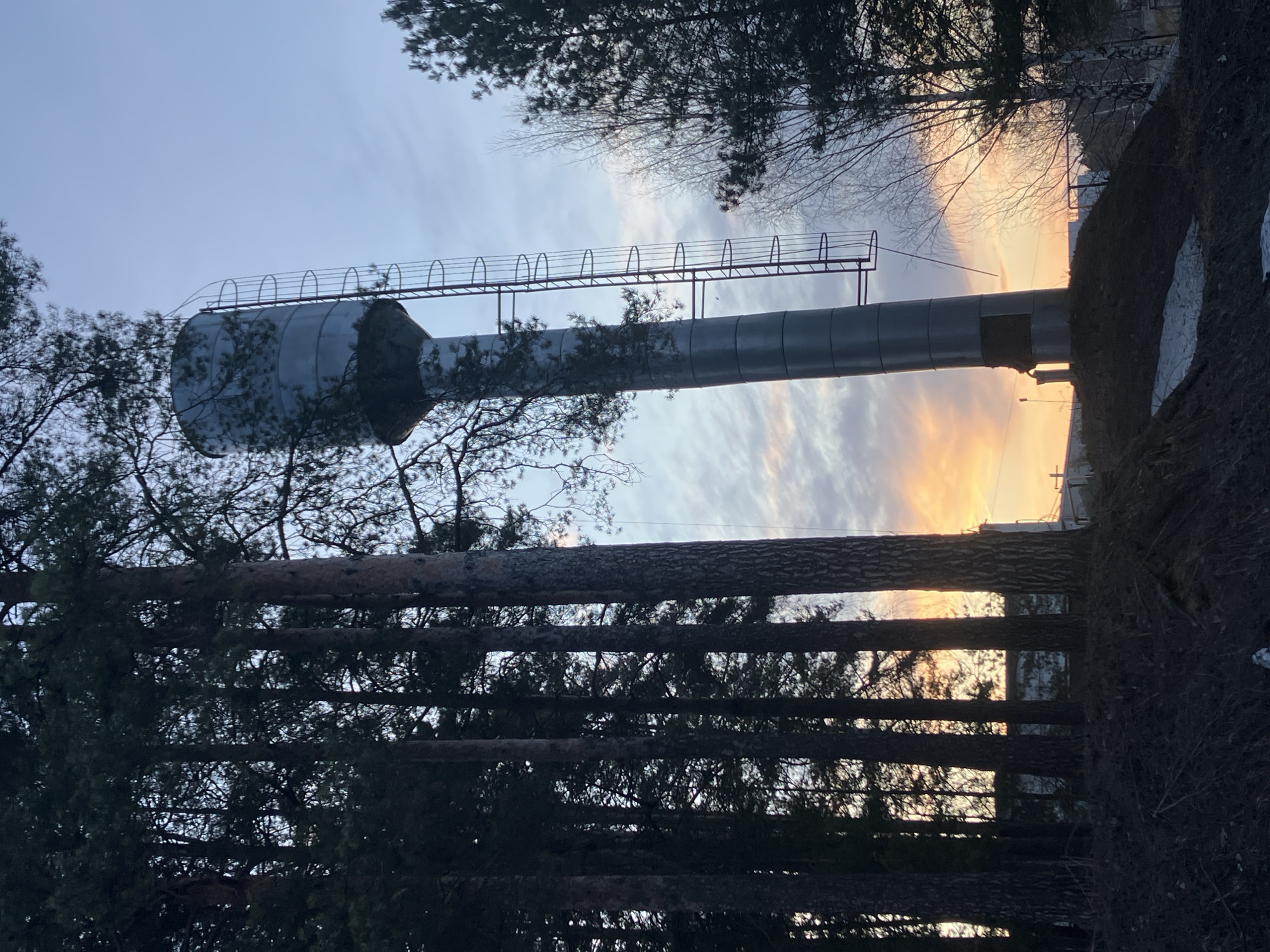 A vertical photograph shows a water tower in the city of Noviy, Russia, surrounded by trees. The water tower is gray and has a ladder and platform near the top. The sky is visible behind the water tower, with orange and yellow hues suggesting either sunrise or sunset. The foreground is dominated by the silhouettes of tree trunks, while the background has more trees and some visible structures. The image is slightly rotated. Snow is seen in the foreground.