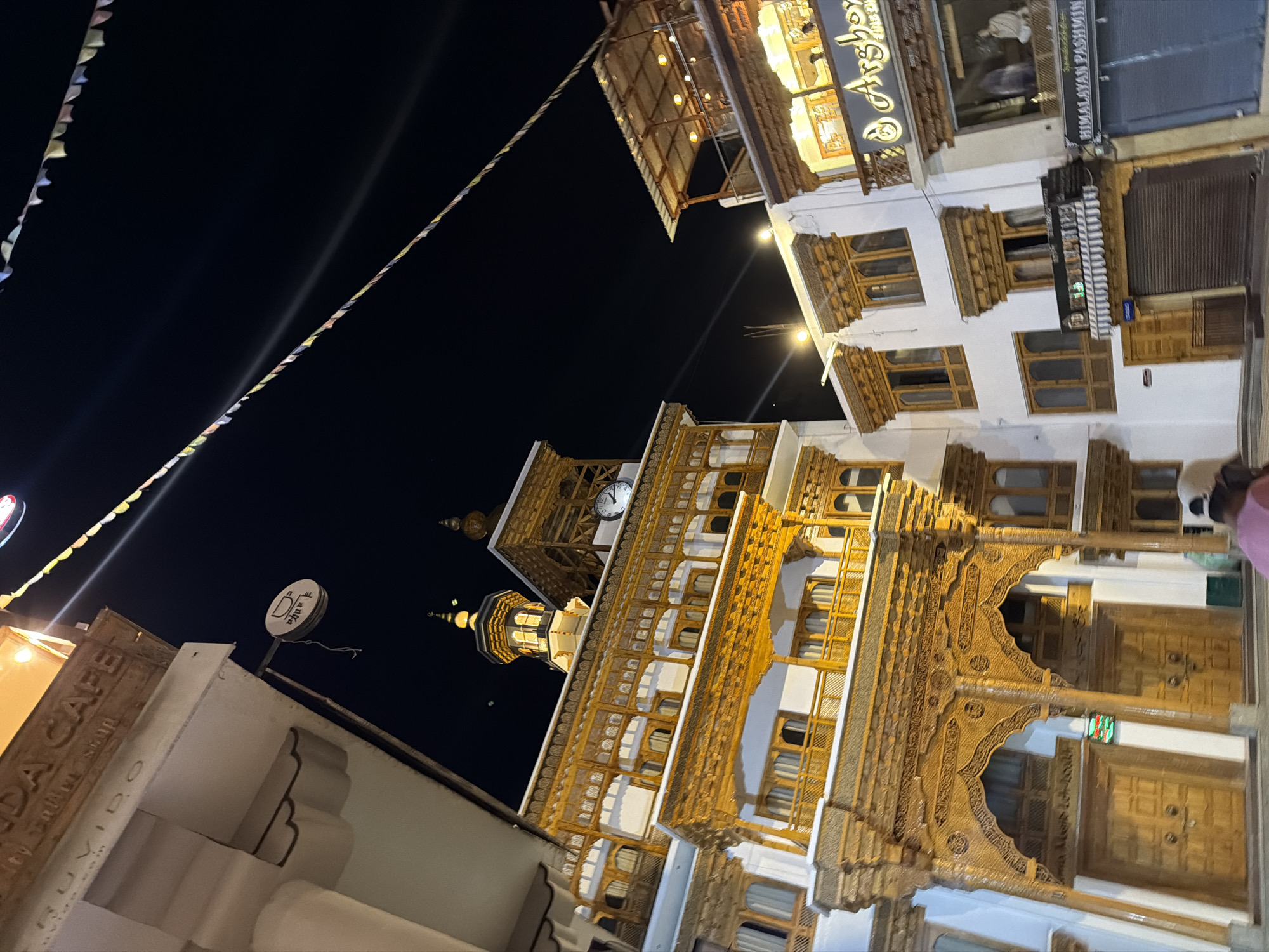 The image captures a night scene in Leh, India, focusing on a street lined with traditional Ladakhi architecture. The buildings feature intricate wooden carvings on their facades and balconies, with multiple stories and large windows. Prayer flags are strung across the dark night sky, adding a touch of color and cultural context.
On the left side of the image, a building has a sign that partially reads "RUVIDO CAFE." A circular sign with a coffee cup symbol hangs from a pole. Further to the right, another building displays a clock face prominently on its facade, showing the time to be around 12:25. This structure also has a dome and minaret, suggesting it might be a mosque or have religious significance.
In the foreground and to the right, a shop sign reads "Arshoy." Below this, another sign indicates "HIMALAYAN PASHMINA." The shops appear to be open or recently closed, with lighting visible inside. A closed shutter is visible on a shop below.
In the bottom right corner, a person wearing a pink garment is partially visible, facing away from the camera. Another figure in white is seen through a window of the "Arshoy" shop. The street appears to be paved, and the overall atmosphere is calm and illuminated by the building lights and the night sky. The architectural style suggests this is a prominent street in Leh's bazaar area.