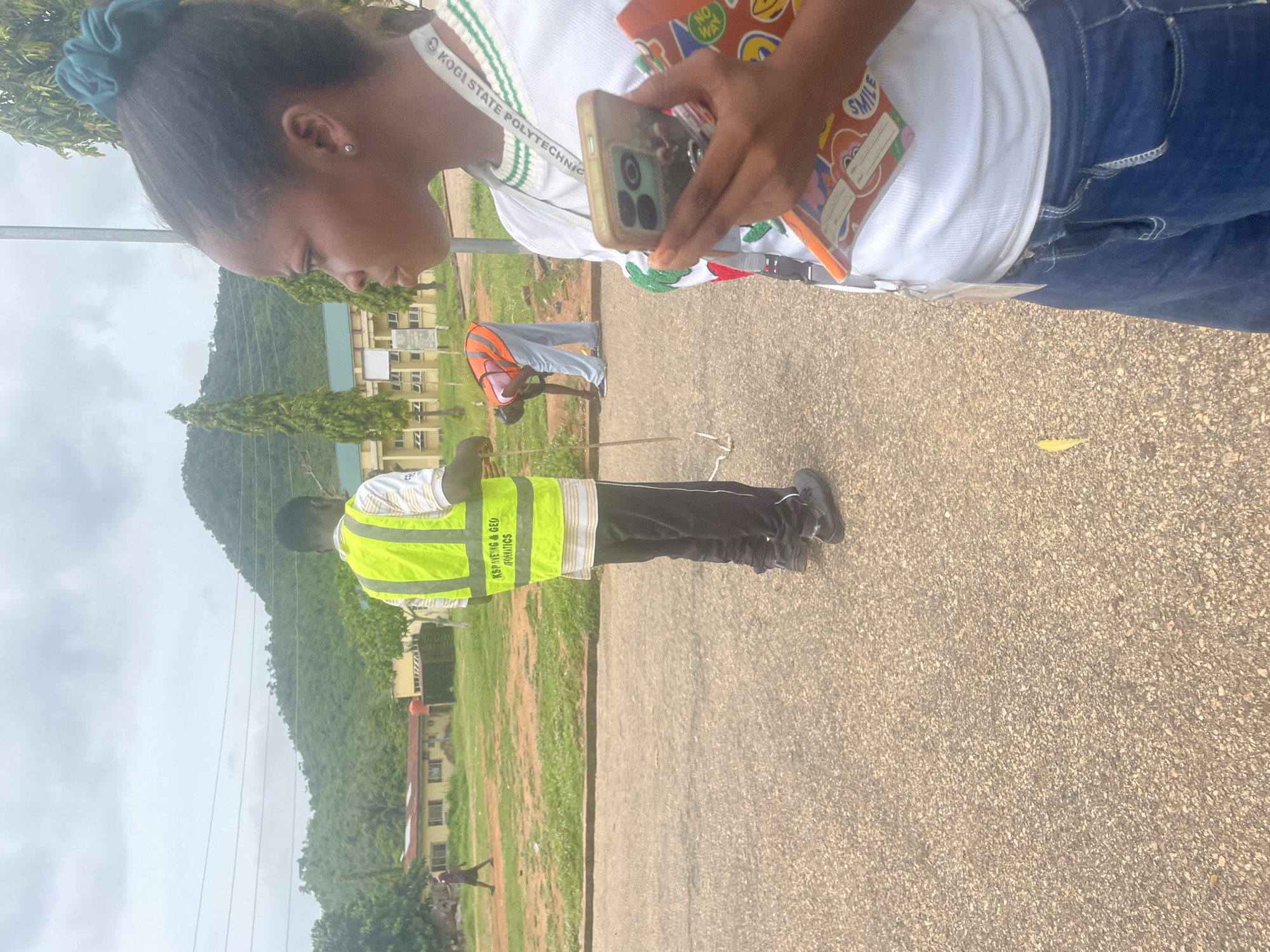 In Lokoja, Nigeria, a young woman is in the foreground, her profile turned to the right, holding a smartphone and an open notebook with a colorful cover. She wears a white t-shirt with green trim and blue jeans. Her hair is pulled back with a green scrunchie. Behind her, a man in a bright yellow reflective vest is visible, holding a measuring tape. He wears black pants and black shoes. Further in the background, another person in an orange vest is bending down, possibly working on the ground. The scene appears to be on a paved road with a grassy area and buildings in the distance, set against a backdrop of lush, green hills under a cloudy sky. The atmosphere suggests an outdoor educational or survey activity.