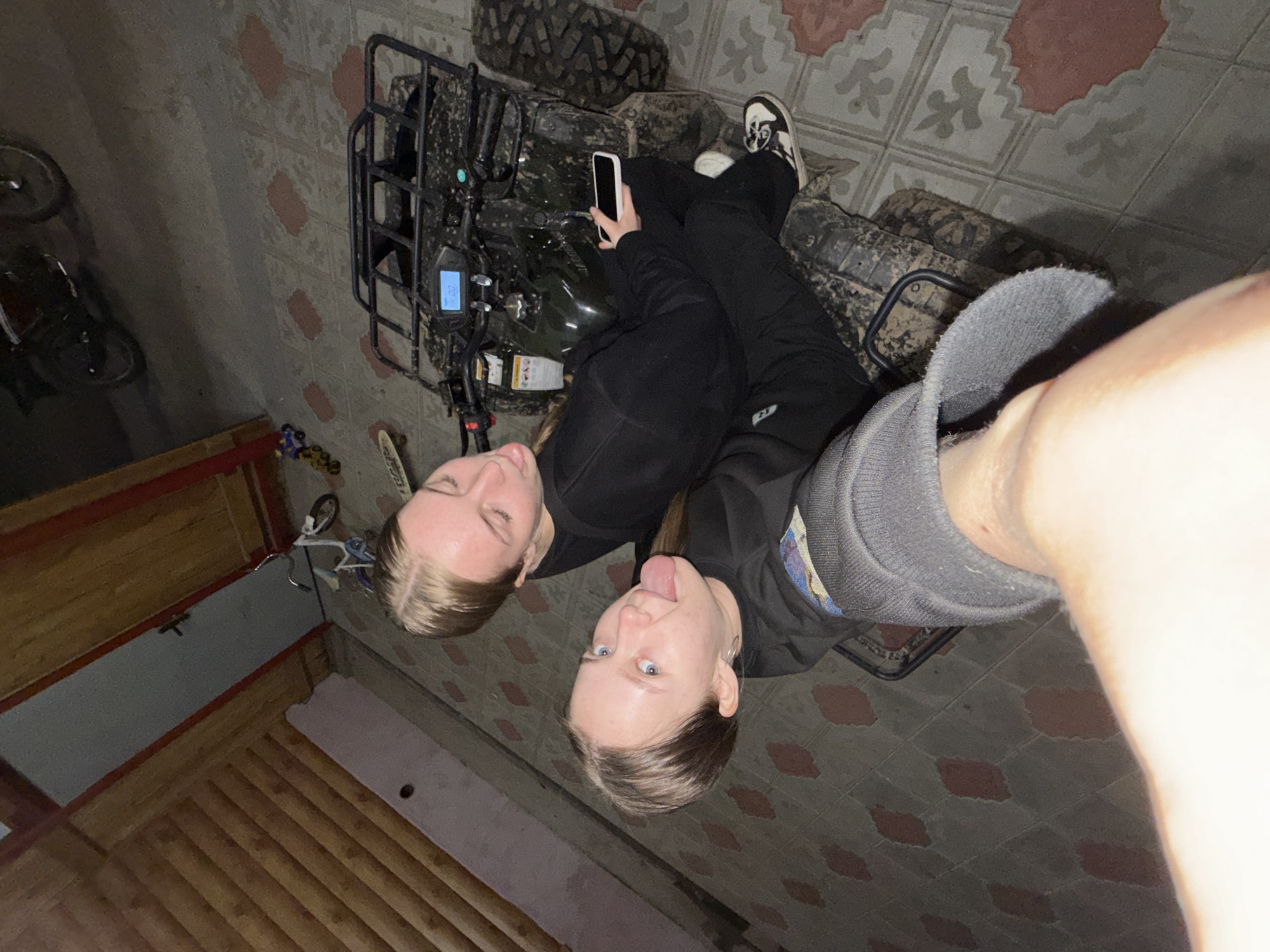 Two fair-skinned individuals, likely female, are positioned above and to the right of a black all-terrain vehicle (ATV). The individual closer to the camera extends their tongue. One individual holds a mobile phone. The ATV appears muddy. The background consists of tiled flooring with a patterned design and wooden planks. A bicycle is visible in the lower left. The location is Zhigalki, Russia.