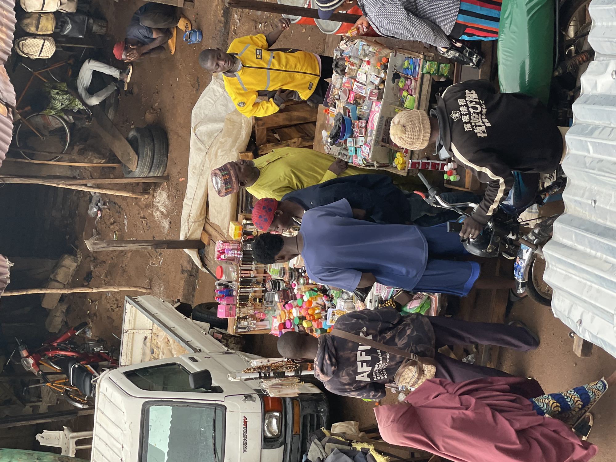 The image captures a bustling street scene in Bukuru, Nigeria. A white truck is parked in the foreground, partially obstructing a view of a storefront. Several people are gathered around a vendor's stand laden with various items, including cosmetics and accessories. The customers are dressed in a mix of casual and traditional attire. A man in a yellow jacket stands out among the crowd. A motorcycle parked on the edge of the frame indicates a common mode of transportation. The background reveals a more cluttered area, with a bicycle, tires, and miscellaneous objects stored under a makeshift shelter. The overall impression is a lively commercial environment.
