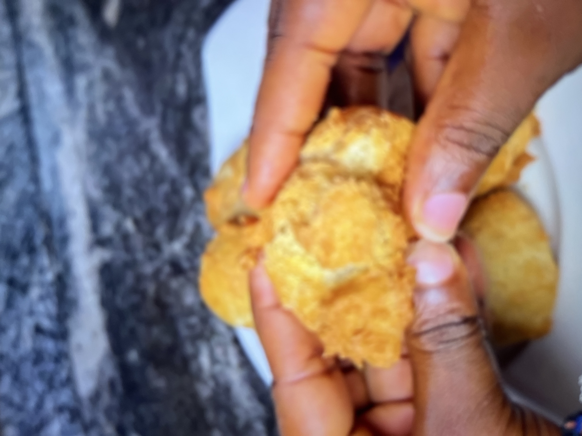 A person is holding a golden-brown, fried pastry in their hands. The pastry appears to be crispy and has a somewhat irregular shape, with a textured exterior. Two hands are visible, one on each side of the pastry, breaking it apart to reveal the fluffy interior. Several other similar pastries are visible on a white plate in the background. The background is blurred but appears to be a countertop with a dark, marbled pattern, possibly granite, which is a common material used in kitchens and countertops in Abuja, Nigeria. The lighting suggests an indoor setting, likely a home kitchen.
