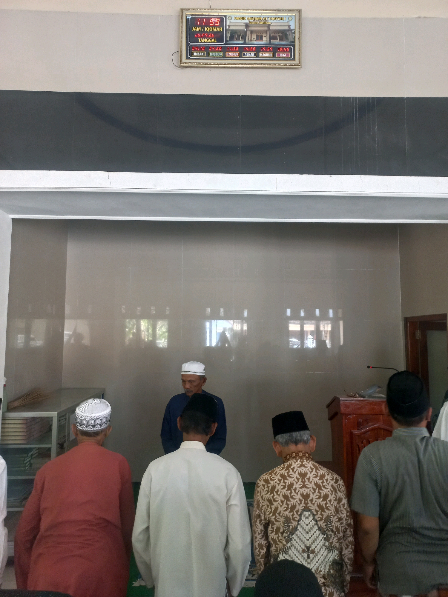 The image depicts an interior view of a mosque, likely located in Nguntoronadi, Indonesia, as indicated by a digital clock displaying prayer times for the local area. The clock is mounted on a wall and shows the time as "11 59" and the date as "JU,PRN,dt" followed by prayer times such as "IMSAK", "SUBUH", "DZUHUR", "ASHAR", "MAGHRIB", and "ISYA". Below the clock, the mosque's name is partially visible as "MASJID SUN...THAYIB...".
In the foreground, several men are standing, facing away from the camera, seemingly preparing for or in the midst of prayer. They are wearing traditional Islamic attire, including caps and long-sleeved shirts. One man in the center, slightly behind the others, is wearing a white cap and a dark blue shirt. To his right, a wooden podium is visible, with a microphone attached.
The walls of the mosque are tiled with a neutral-colored material, and windows with blinds can be seen in the background, reflecting light and glimpses of the outdoors. To the left, a metal shelf unit holds stacks of books, and a broom leans against it. The lighting appears to be artificial, supplemented by natural light filtering through the windows.
