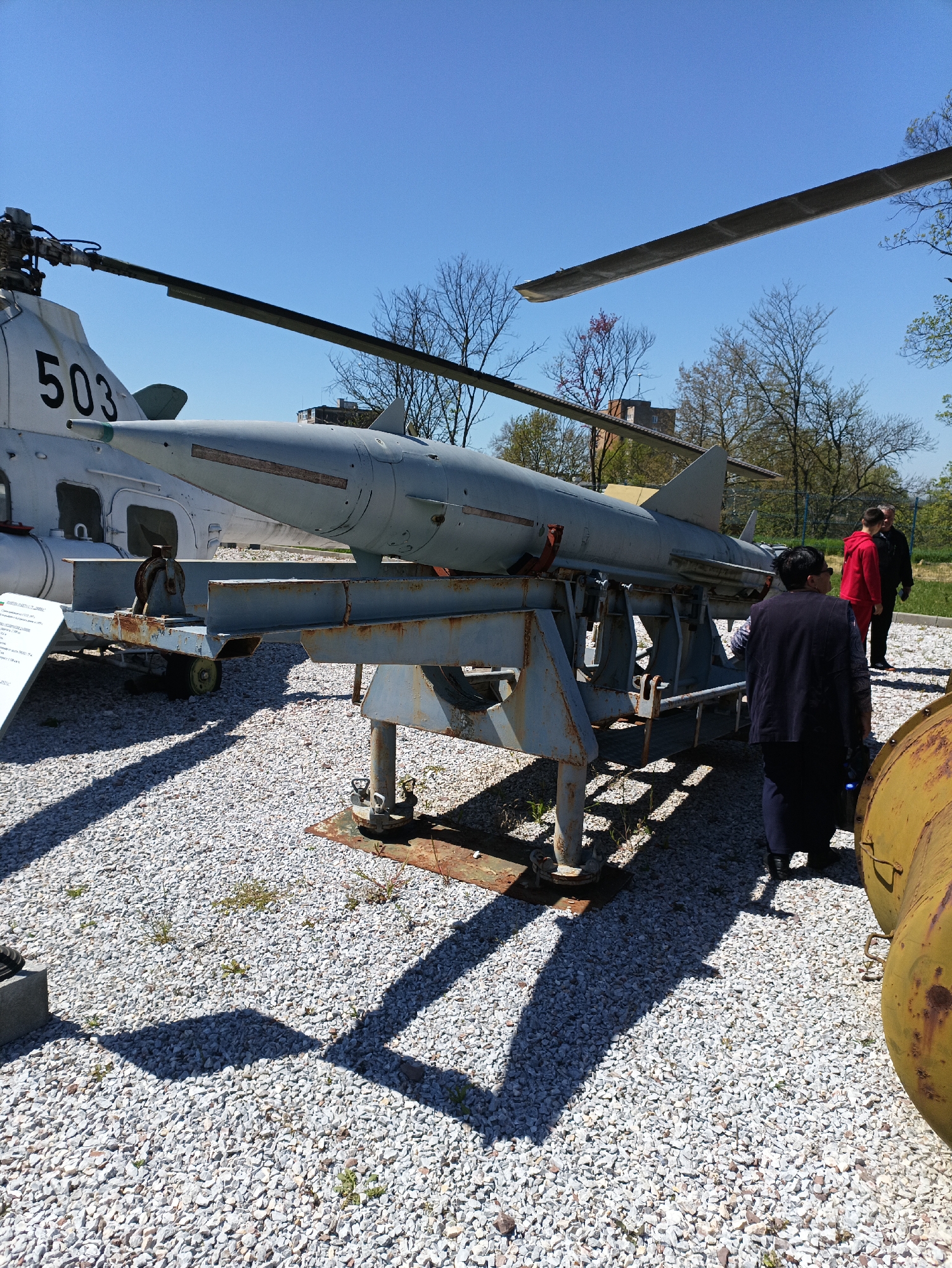 The image depicts a static display at the Yambol Military Museum in Yambol, Bulgaria. The main subjects are a gray missile mounted on a weathered, bluish-gray metal trailer, and a helicopter marked with the number "503" on its tail visible in the background. The ground is covered in small white gravel.
There are three people in the scene. One person is wearing a dark blue jacket and standing near the missile trailer. Two people, one wearing a red jacket and the other wearing a black jacket, stand together further in the background.
Other objects present include a rusty yellow cylindrical object near the lower right corner, tree branches visible in the background, and building in the background.
The environment appears to be outdoors on a clear, sunny day, casting distinct shadows from the objects. The overall scene suggests an outdoor military exhibition.