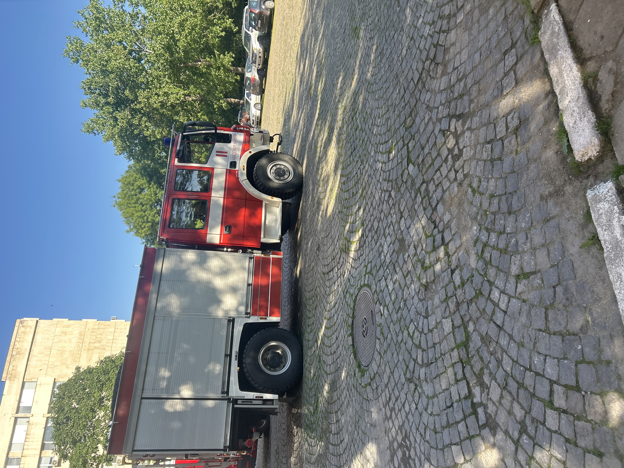 The image depicts a red and white fire truck parked on a cobblestone street in Sofia, Bulgaria. A building is visible in the background with multiple parked cars next to the fire truck. There are trees in the image as well. The scene is outside in daylight with clear blue sky. A manhole cover can be seen on the cobblestone street.