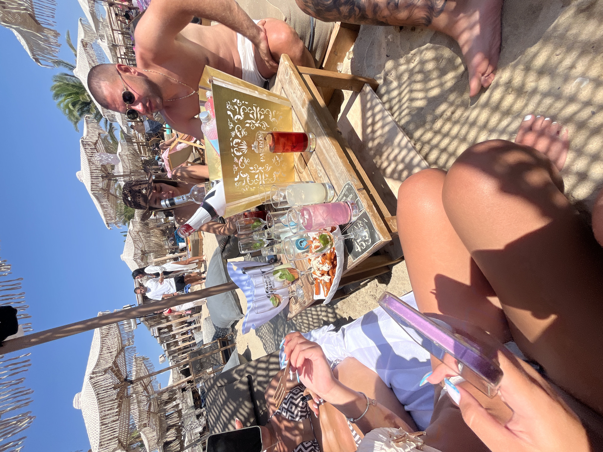 The image depicts a sun-drenched beach scene in Néa Irakleia, Greece, likely a beach club. Several people are present, relaxing under a canopy of large, macrame-fringed umbrellas. In the foreground, a group of individuals are seated around a low wooden table laden with drinks and food.
On the table, a large gold ice bucket, embossed with a decorative pattern and the word "PATRON", contains several bottles, one appearing to be a champagne bottle. Surrounding the ice bucket are various glasses, some filled with colorful beverages, including a deep red drink in a tall glass and two glasses containing pink and clear liquids. A plate of fried food, possibly french fries with a topping, is also on the table, along with a sprig of greenery and some cucumber slices.
A person on the right, a man, is wearing sunglasses and white swim trunks, his torso bare. To his left, a woman is seated, wearing a dark hat and a bikini top. Another person, partially visible in the foreground, is holding a phone and a translucent purple object. In the middle ground, a couple stands near the water's edge, the man in a white t-shirt and black shorts, the woman in a white cover-up over a bikini. The beach itself is sandy, with beach chairs and loungers scattered throughout. The overall atmosphere suggests a leisurely day spent by the sea.