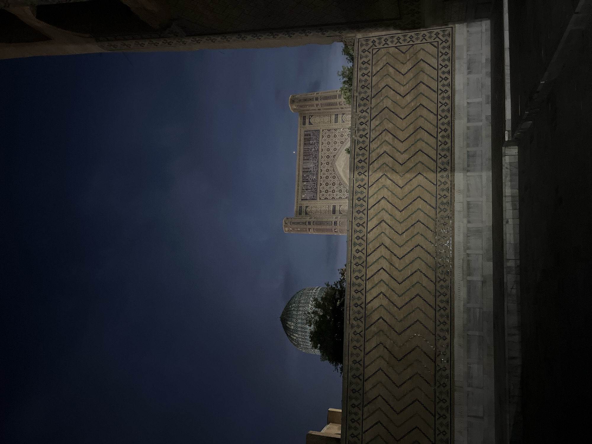 This media file captures a nighttime view of an architectural complex in Samarkand, Uzbekistan. The scene is dominated by the dark night sky, with a sliver of the crescent moon visible near the top of the central building. The architecture features intricate geometric patterns and mosaic tilework, characteristic of Islamic art and architecture.
The foreground is partially obscured by a dark, shadowed structure on the right, creating a framing effect. To the left, a portion of the sky reveals a towering, ornate building, possibly a mosque or madrasah, with decorative facades. Below this, a prominent dome, likely belonging to a mausoleum or another significant structure, is visible, partially enveloped by greenery. The detailed tilework on the exterior of the buildings is a striking element, even in the low light. The overall impression is one of grandeur and historical significance, enhanced by the serene and contemplative atmosphere of the night. The image suggests a peaceful and quiet evening, likely in a historical or religious site. There is no visible text in the media file.