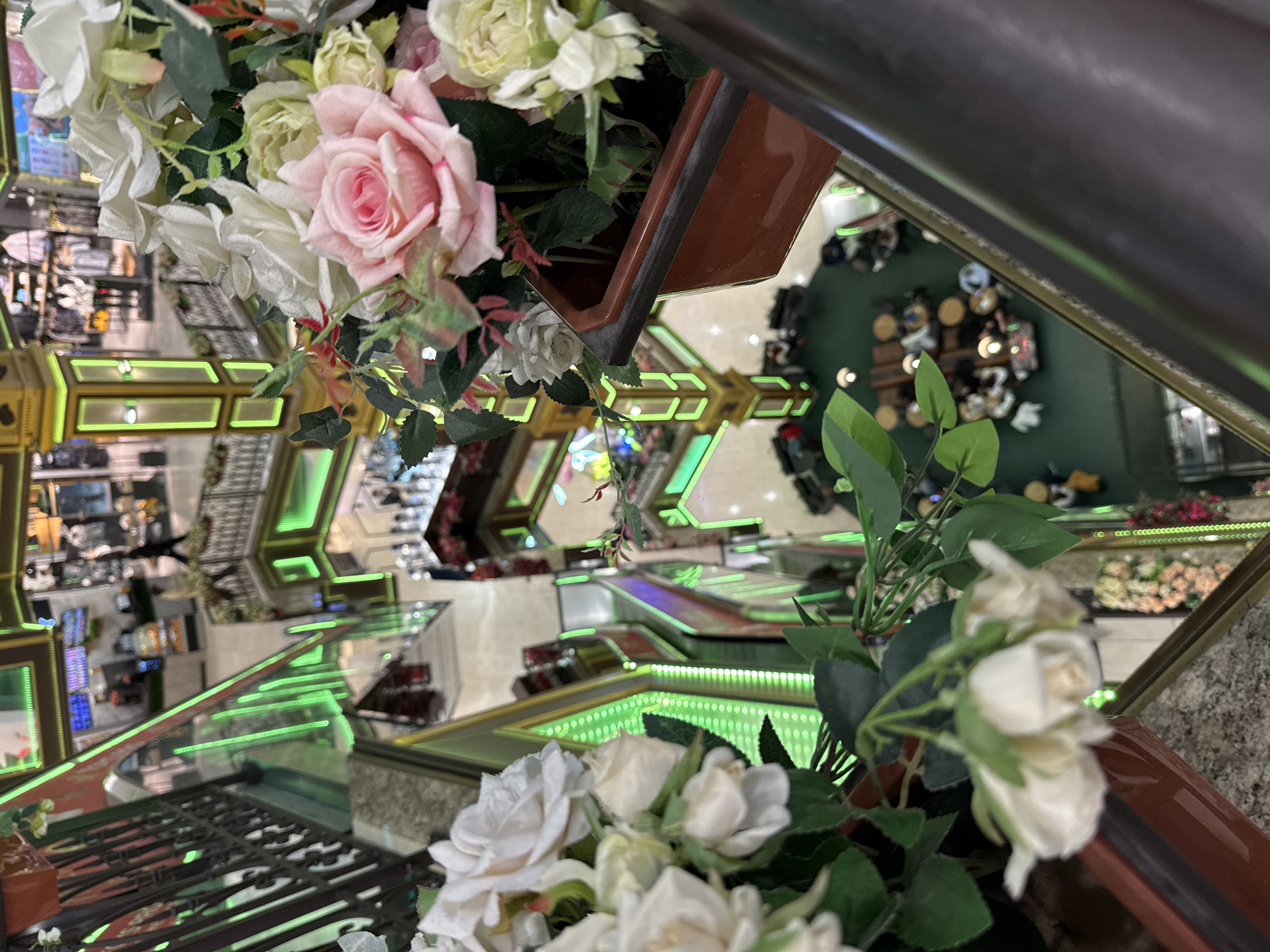 From an elevated vantage point in Moscow, Russia, the image presents a view through a railing adorned with floral arrangements in pots. The flowers are primarily white and pink roses, with green foliage. Below, an interior space featuring escalators illuminated with green lighting is visible. The reflection on the glass surfaces creates a mirrored effect, distorting the view. In the lower right corner, a green-carpeted area with tables and chairs suggests a dining or seating space, occupied by several individuals. The architectural design includes geometric shapes and a multi-level layout. The overall scene gives the impression of an upscale shopping mall or commercial building.