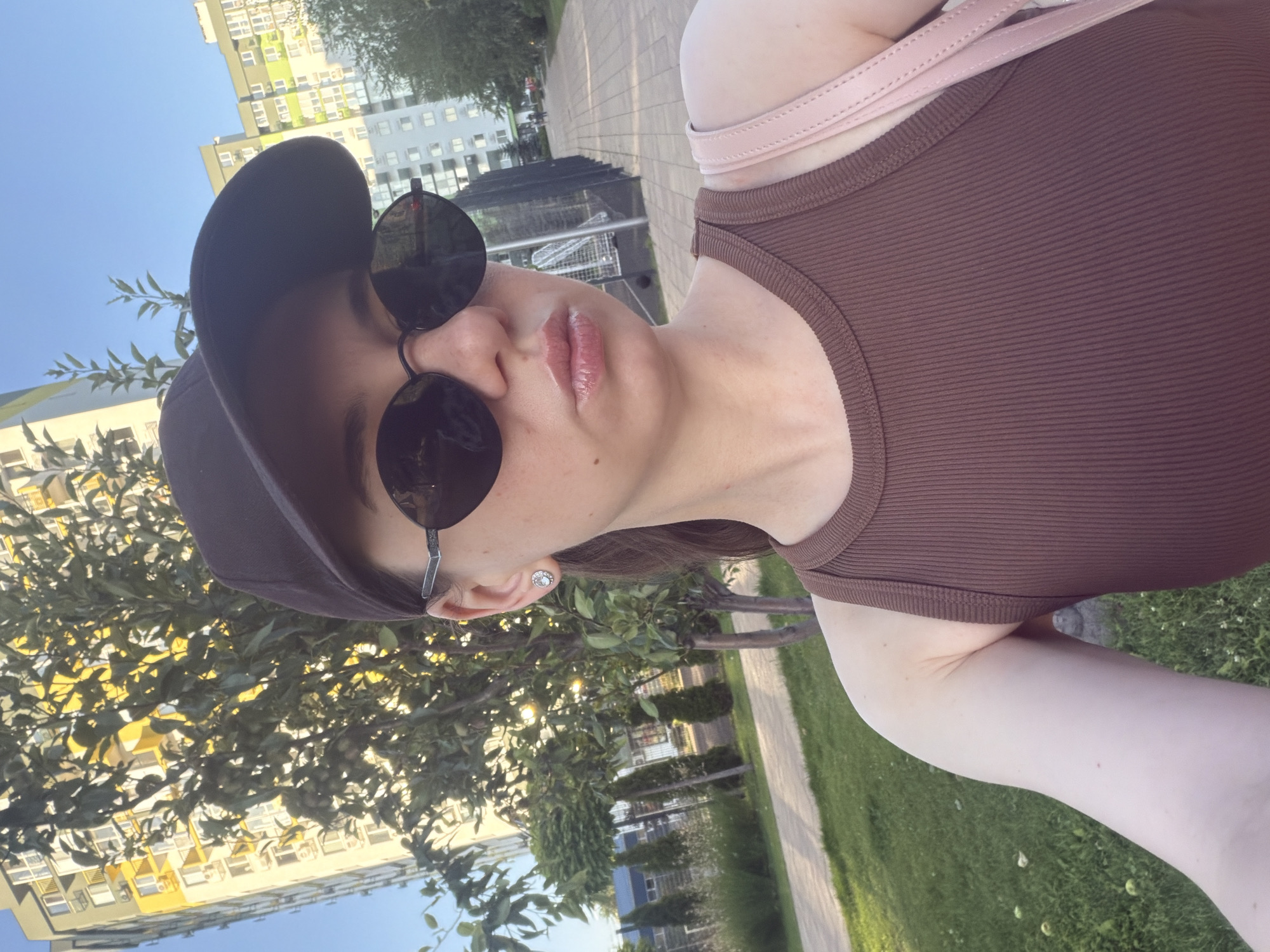 A young woman, wearing a brown ribbed tank top, black baseball cap, and oval sunglasses, is taking a selfie outdoors. She has light skin, brown hair pulled back, and her lips are glossy. She is also wearing a diamond stud earring. The background shows a modern apartment building with yellow and white facade, a green tree with sunlight filtering through its leaves, and a paved walkway. In the distance, another apartment building with green and white facade is visible. A pink strap of a purse or bag is visible over her shoulder. The scene appears to be in a residential area, possibly a park or courtyard, in the city of Kyiv, Ukraine, given the architectural style of the buildings and the presence of manicured trees and greenery.