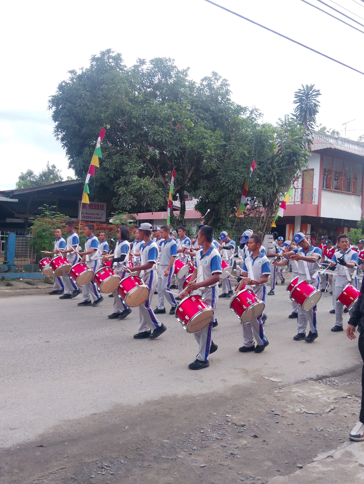 A marching band composed of young males and at least one female is parading down a street in Serbelawan, Indonesia. The participants, dressed in matching blue and white uniforms with grey pants, are playing red bass drums. They are holding drumsticks and marching in formation. In the background, there are trees, buildings, and Indonesian flags. One building in the background has a sign that reads "UD. MERBAU" and "Jl. Merdeka NO. 17 SERBELAWAN". The street is paved with asphalt, and there are some rocks and debris on the side of the road. The sky is overcast.