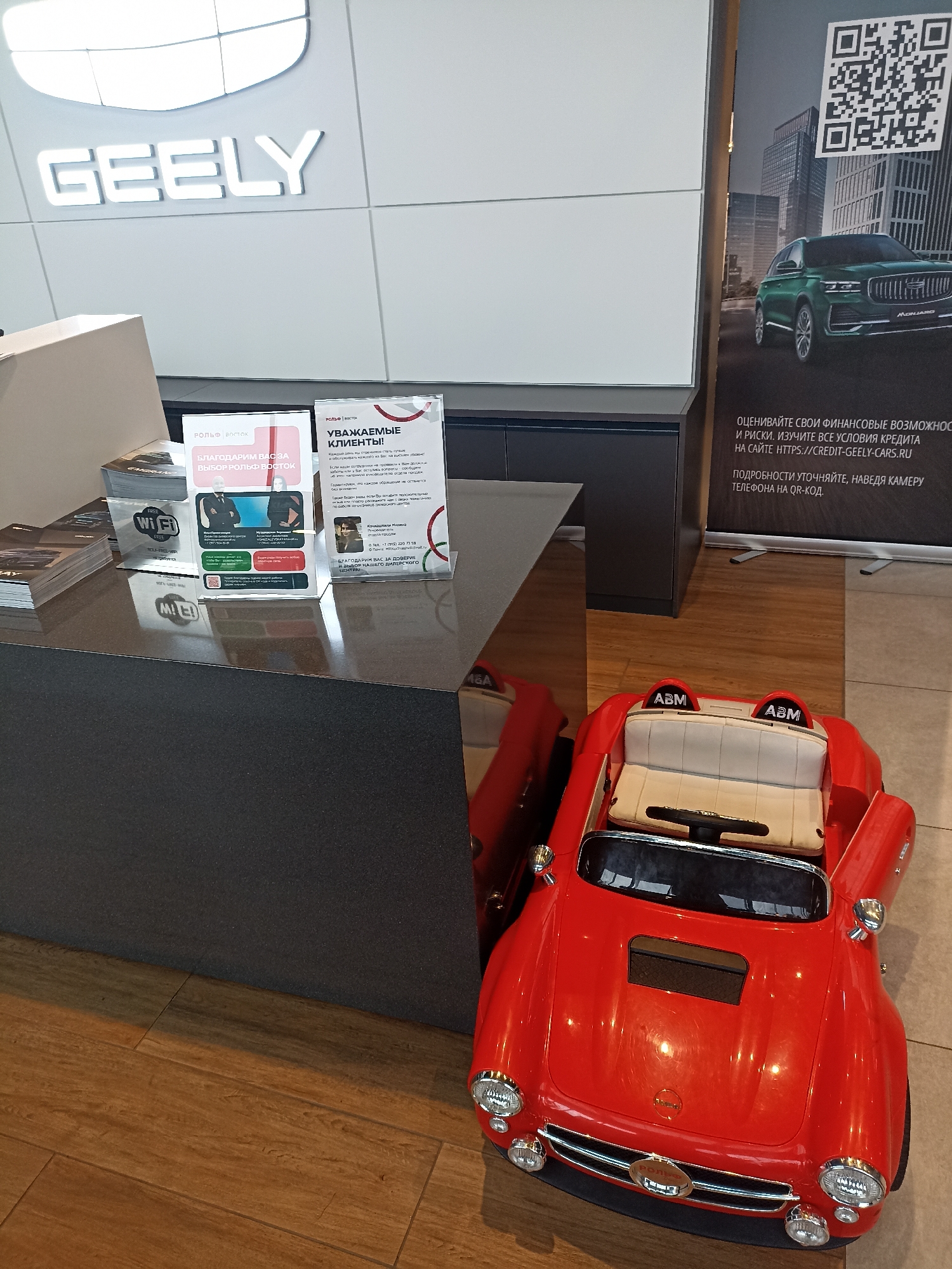 The image depicts the interior of a Geely car dealership in Moscow, Russia. The dealership features a prominent "GEELY" logo illuminated on a white wall in the background. In the foreground, a dark gray reception desk is visible, with various brochures and informational stands placed on its surface. One stand, in pink and white, thanks customers for choosing Geely. Another stand, also with Russian text, has a QR code and text related to financing options. To the right of the desk, a large banner displays a green Geely SUV and a QR code, with further text in Russian about financing. On the floor, a red, miniature replica of a car, likely a toy for children, is positioned next to the reception desk. The environment appears to be a modern, well-lit showroom with wooden flooring. No people are clearly visible in the scene.