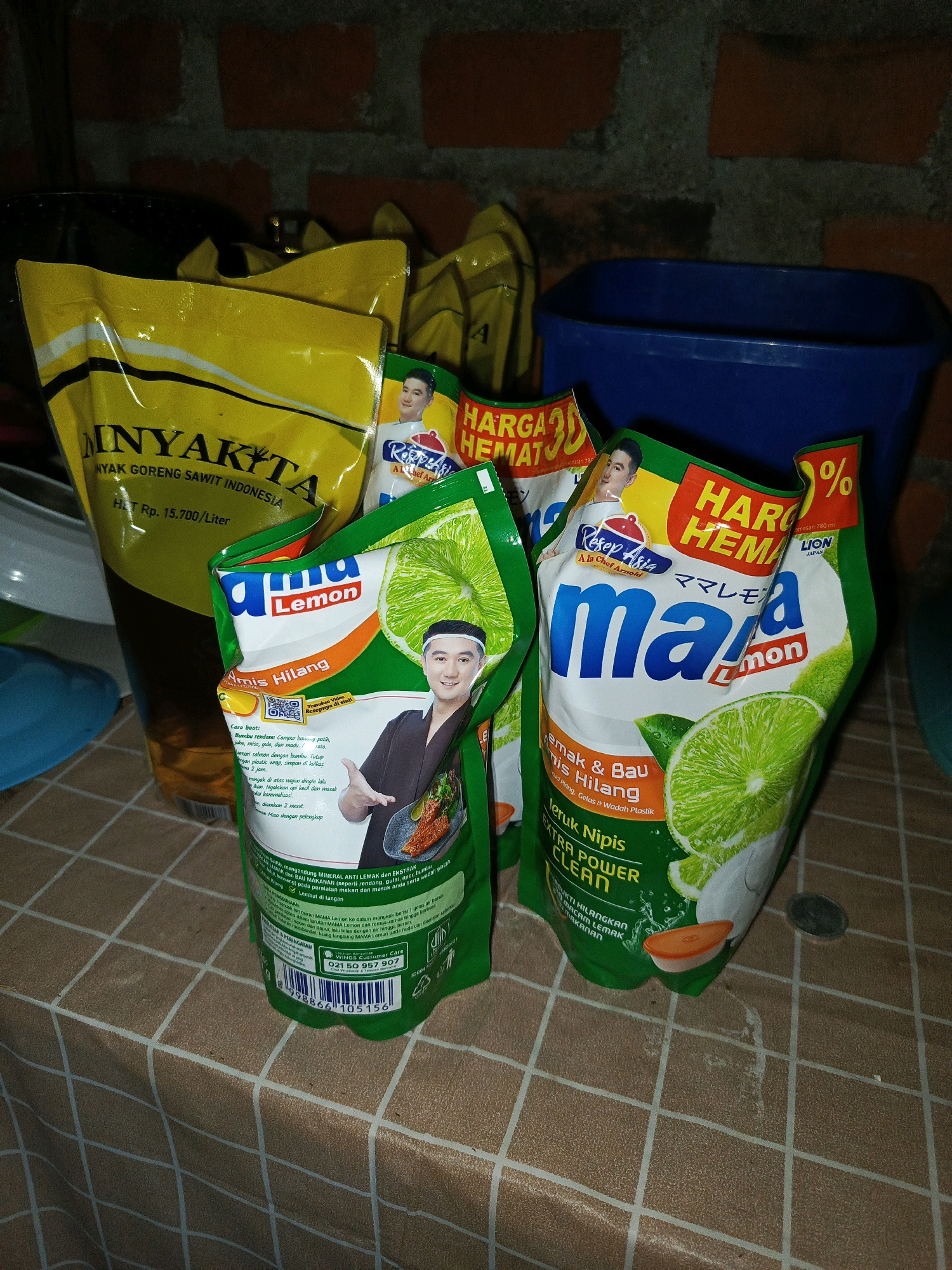 A close-up, slightly blurry, indoor shot captures a collection of household cleaning products and cooking oil arranged on a checkered tablecloth. In the foreground, two green pouches of "MAMA Lemon" dish soap with lime imagery and graphics of limes are prominently displayed. One pouch is more visible than the other, showing text related to cleaning power. Behind these, a large yellow pouch of "Minyakita" cooking oil is partially visible, with text indicating it is "Minyak Goreng Sawit Indonesia." A dark blue plastic container is in the background, and a brick wall is visible at the top of the frame. The overall scene suggests a domestic setting, possibly a kitchen counter or dining table. The products and their labels are the primary subjects of the image, with no people or discernible actions taking place. The location is not evident from the image itself.