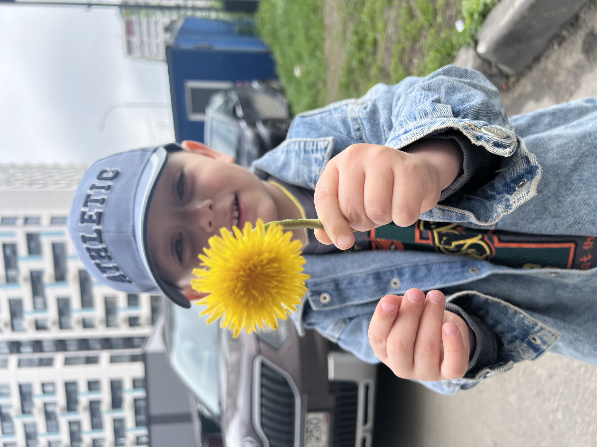 A young boy stands outdoors, holding a yellow dandelion. He wears a denim jacket and a blue baseball cap with "ATHLETIC" printed on it. The boy is smiling. In the background are buildings and cars, indicating an urban environment. The presence of cars with Russian license plates suggests the location could be Mytishchi, Russia. The setting appears to be on a sidewalk or near a street.