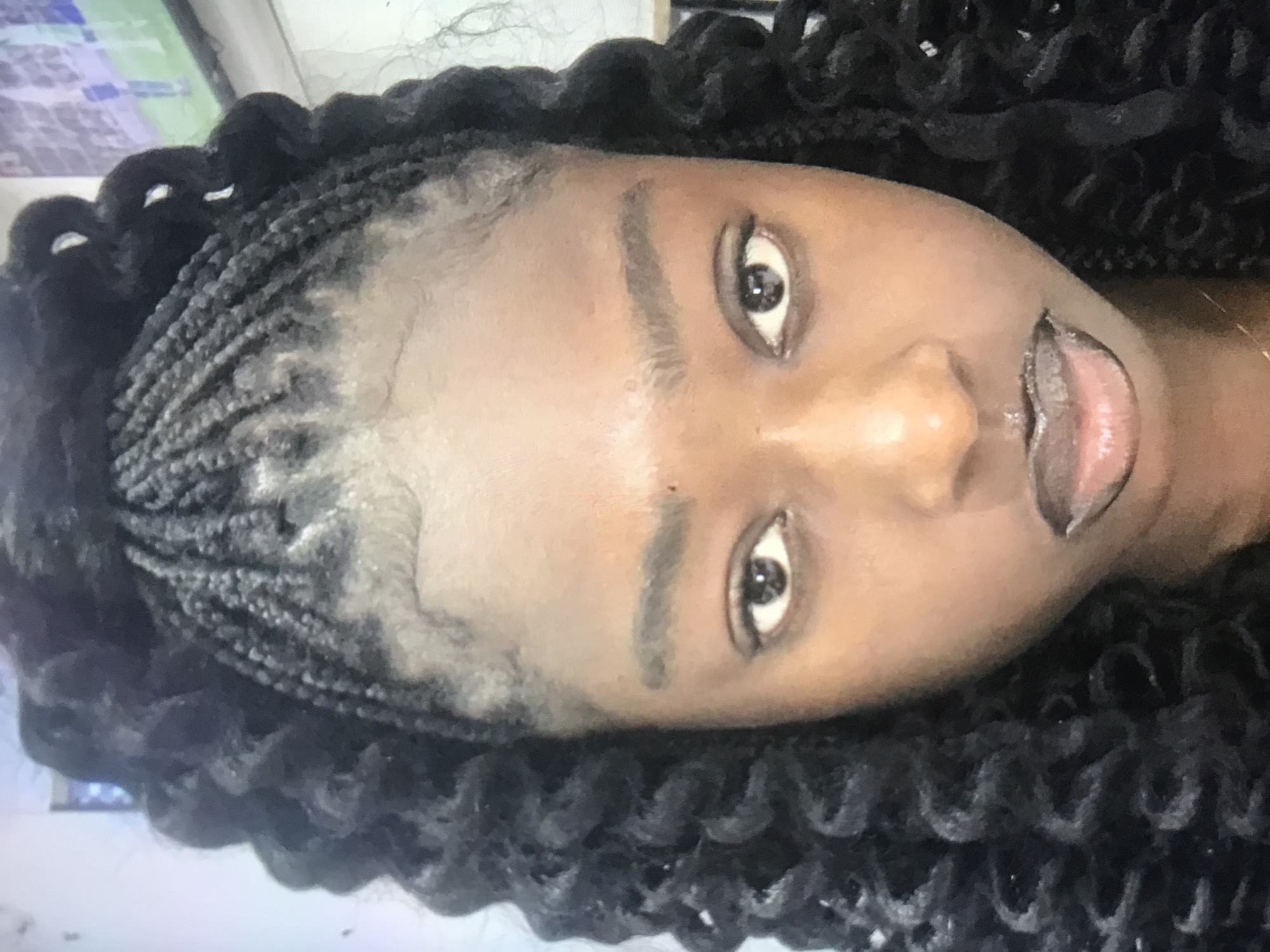 A medium-shot photograph shows a woman with a dark complexion facing the camera. Her hair is styled in neat cornrow braids at the front, transitioning into loose, curly extensions at the back. She has dark eyes and prominent eyebrows, and she is wearing lipstick with a dark outline. The image suggests the scene is indoors. There are no indications of the location of Unguwar Saya, Nigeria.