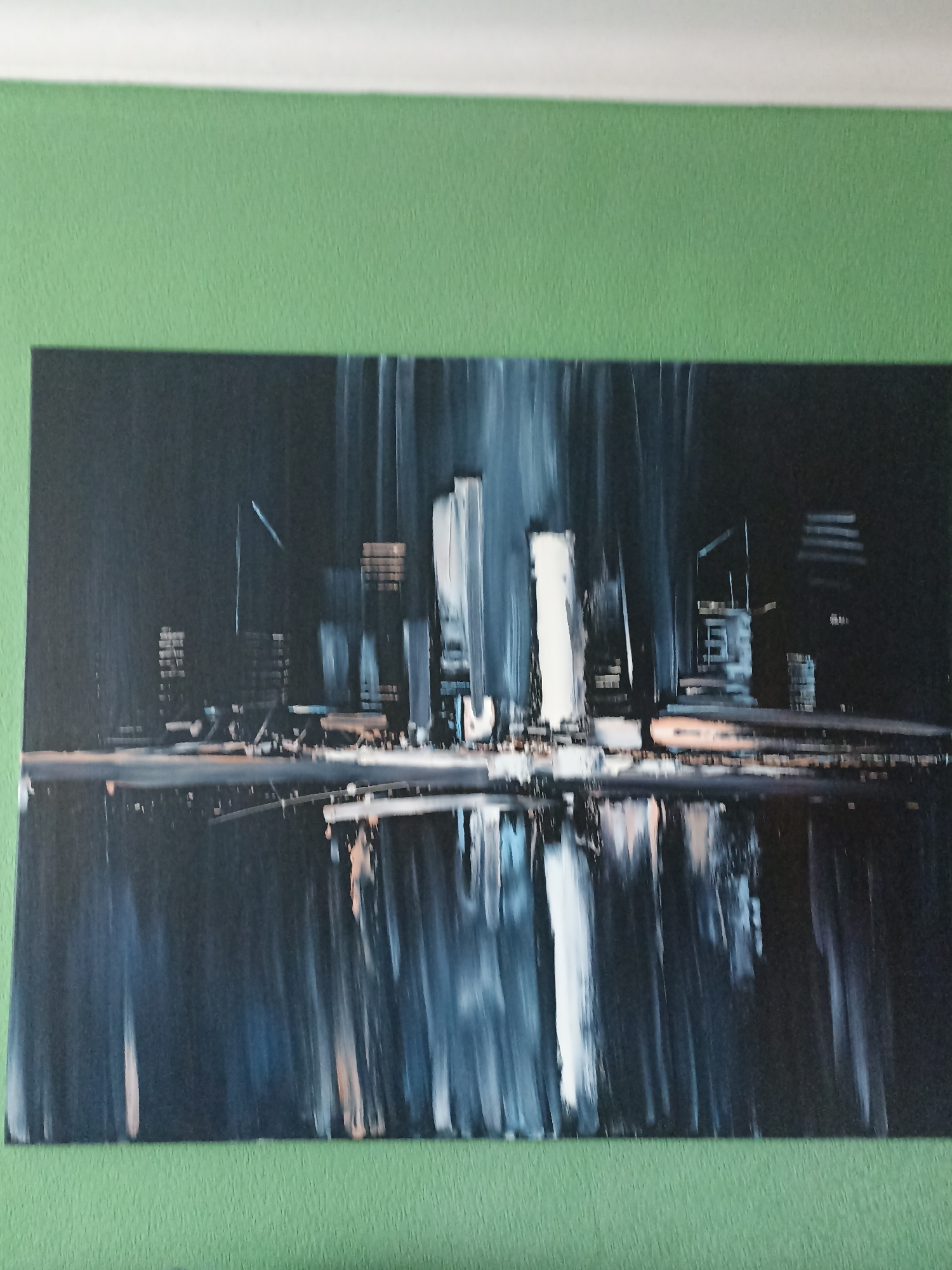 The image shows an abstract painting of a cityscape with a dark, moody atmosphere. The painting depicts tall, vertical structures that suggest skyscrapers, with various shades of gray, black, and white used to create a sense of depth and illumination. Reflections of the city are visible in the water below, adding to the overall ethereal quality of the artwork. The painting is displayed on a textured green wall. There is no indication in the image that this scene is located in Novosibirsk, Russia.