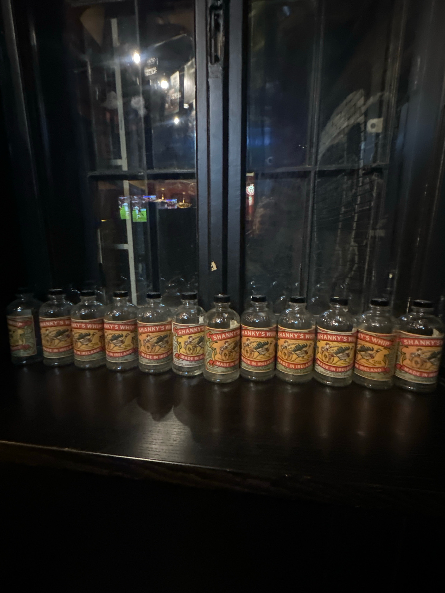 A line of identical miniature bottles with orange and yellow labels sits on a dark wooden surface. The labels, which appear to be from "Shanky's Whip" whiskey, feature a drawing of a cannon. The bottles are clear glass with black caps. Behind the bottles, a dark window or pane of glass reflects light and shows blurry shapes, possibly indicating an indoor setting like a bar or pub in Belgrade, Serbia. The lighting is dim, with some sources of light creating glare on the glass.