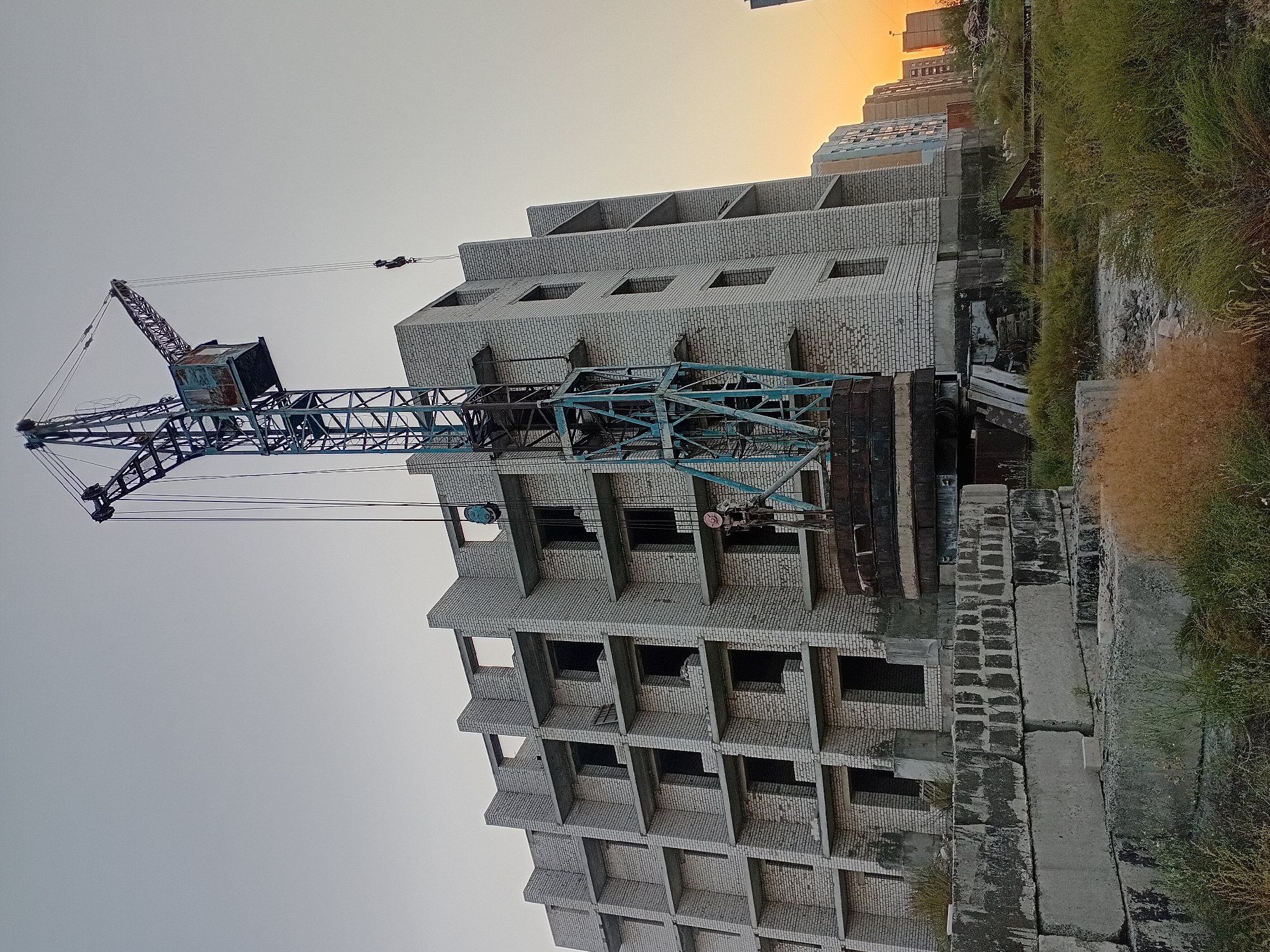 A construction site in Semey, Kazakhstan, features a multi-story building under construction, made of white brick with dark, open window frames. A tall, blue construction crane is positioned next to the building, with its hook and cables extending over the structure. In the foreground, stacks of concrete blocks lie on the ground, interspersed with tall, dry grass. The sky is clear with a warm, orange glow on the horizon, suggesting either sunrise or sunset. Other apartment buildings are visible in the background on the right side of the image. No people are present in the scene.