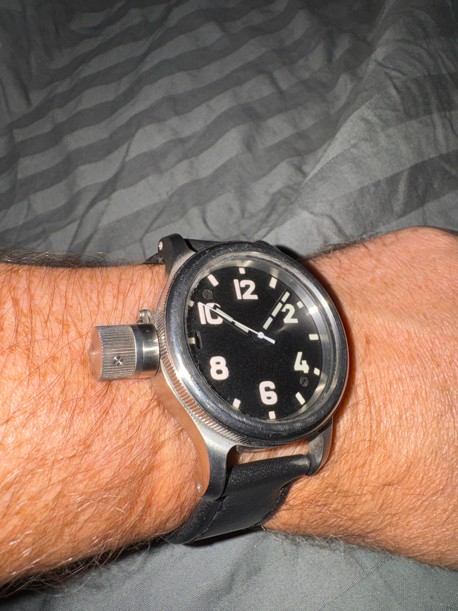 A person's wrist is visible, with a watch on it. The watch has a black face with white numerals and hands. The strap of the watch appears to be black leather. The watch is worn on a hairy arm. The background is a grey, striped fabric, possibly a bedsheet. The image is taken from a close-up perspective, focusing on the watch and wrist. There is no indication of the city of Kommunarka, Russia in this image.