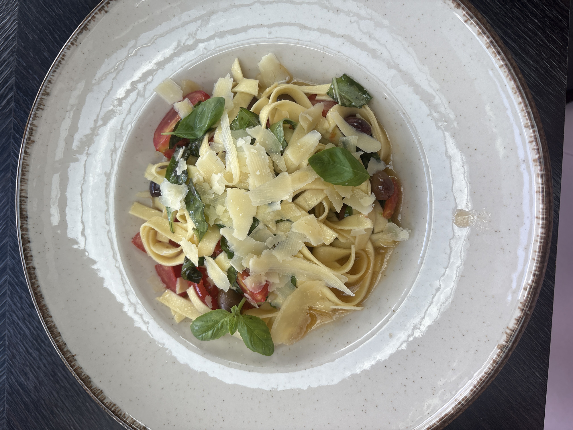 A close-up, top-down shot shows a plate of pasta with cherry tomatoes, olives, basil, and shaved parmesan cheese. The pasta is fettuccine, and it is coated in a light, clear sauce. The plate is white with a speckled finish and a rustic brown rim. The dish is presented on a dark, textured surface, possibly a table. The lighting is natural, casting soft shadows. No people are visible in the image. The location is Unknown, Unknown.