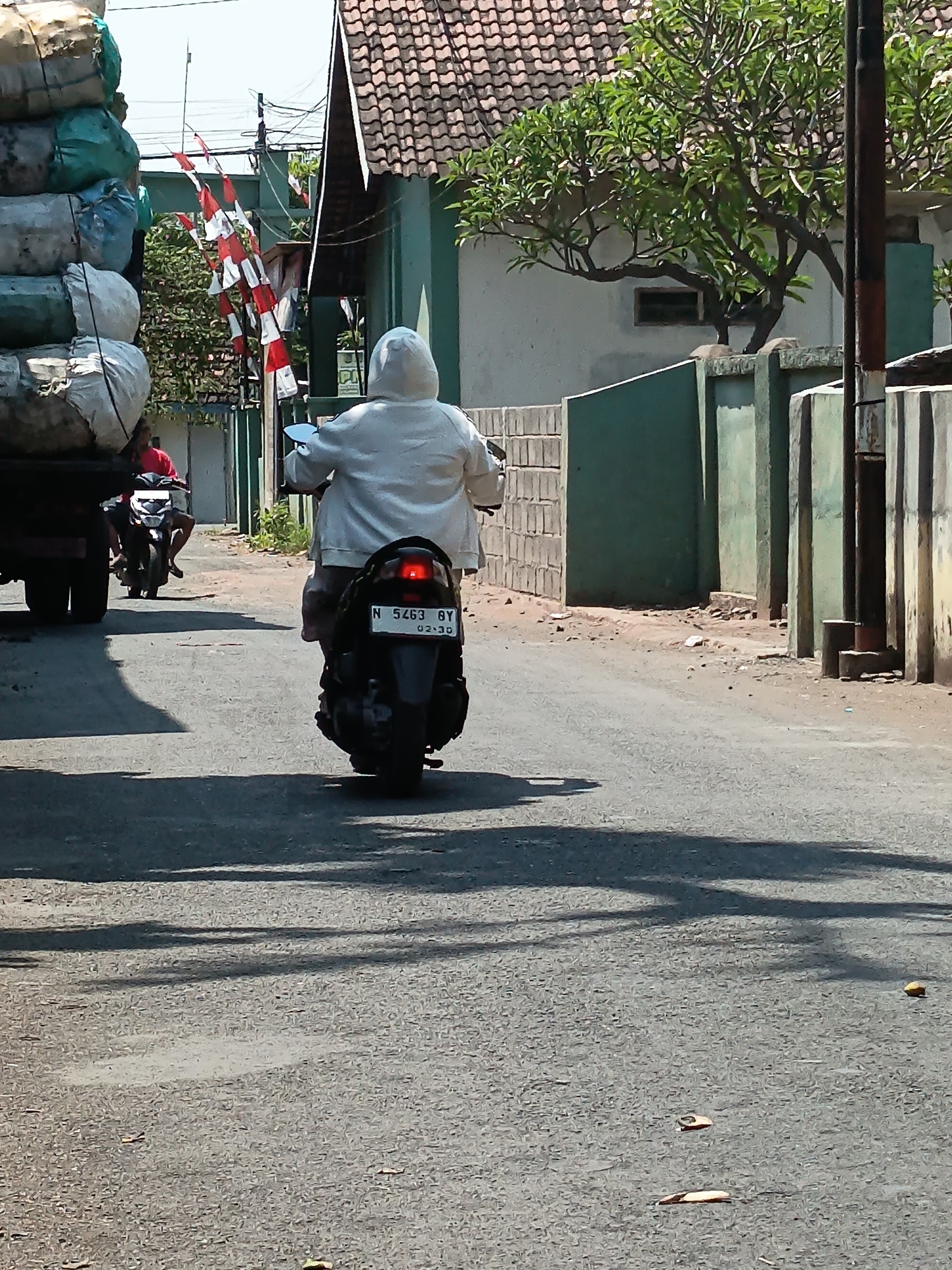 In the village of Kandangjati Wetan, Indonesia, on a sunlit day, a person wearing a white hooded jacket rides a dark-colored scooter down an asphalt road. The scooter has a visible license plate reading "N 5463 BY". In the background, a truck loaded with large white and green sacks is parked on the left side of the road. Further back, another person on a scooter, wearing a red shirt, is visible. Buildings with tiled roofs and green walls line the right side of the road, and a tree with green foliage is situated in front of one of the buildings. Red and white Indonesian flags are strung along a wire, suggesting a local celebration or commemoration. Shadows of trees and possibly the scooter are cast across the road. The environment appears to be a residential or semi-commercial area.
