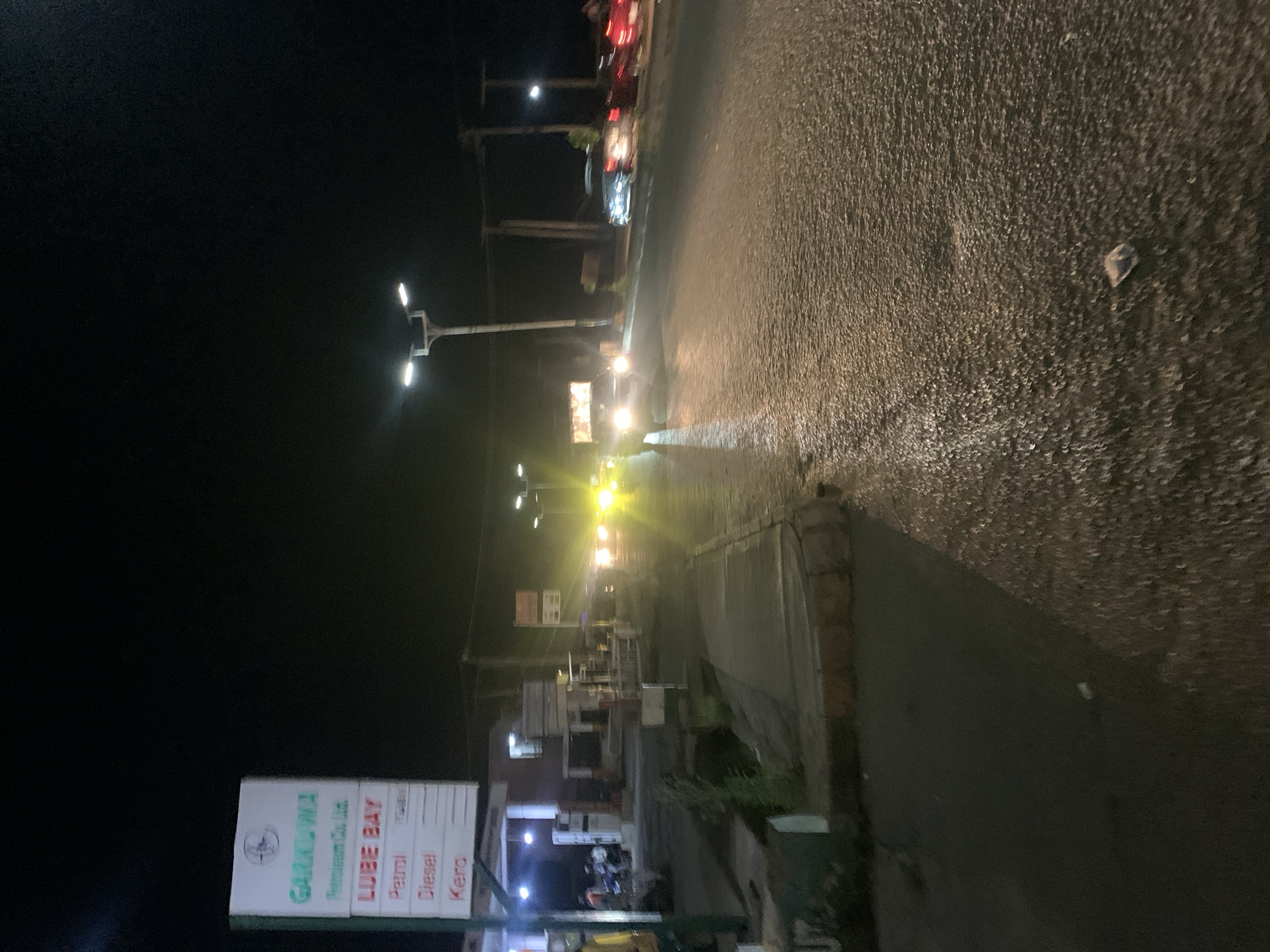 This is a nighttime, low-light, eye-level, slightly blurred, full shot of a street scene in Rafin Par, Nigeria. The image captures a gas station on the left with a sign that reads "GARKOWA Petroleum Co. Ltd. LUBE BAY Petrol Diesel Kero". Several people and motorcycles are visible near the gas station pumps. A street light illuminates the area. The road in front of the gas station is wet and reflects the headlights of passing vehicles. Cars are driving on the road, with their headlights creating bright streaks of light. The environment appears to be a commercial area with buildings and streetlights visible in the background. The overall impression is a busy street at night, with the gas station being a prominent feature.