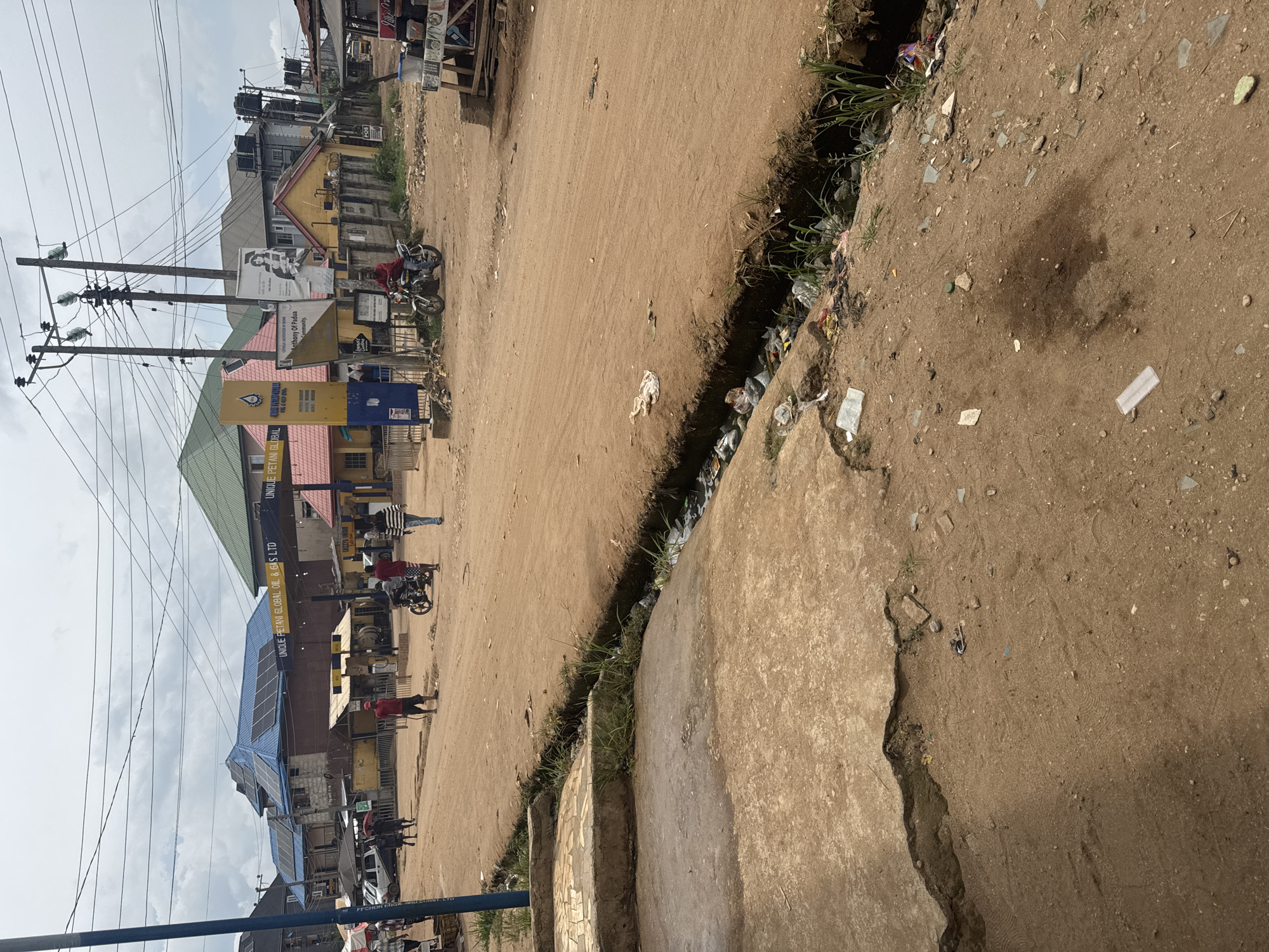 The image depicts a street scene in Ihiagwa, Nigeria. The road is unpaved and made of packed earth. A concrete curb runs along the edge of the road, and a ditch filled with trash is visible between the road and the curb. On the left side of the image, there are buildings, including a gas station with the sign "UNIQUE PETANI GLOBAL OIL & GAS LTD." People and motorcycles can be seen in the area near the gas station. Electrical poles and wires are also visible in the background. The sky is overcast.