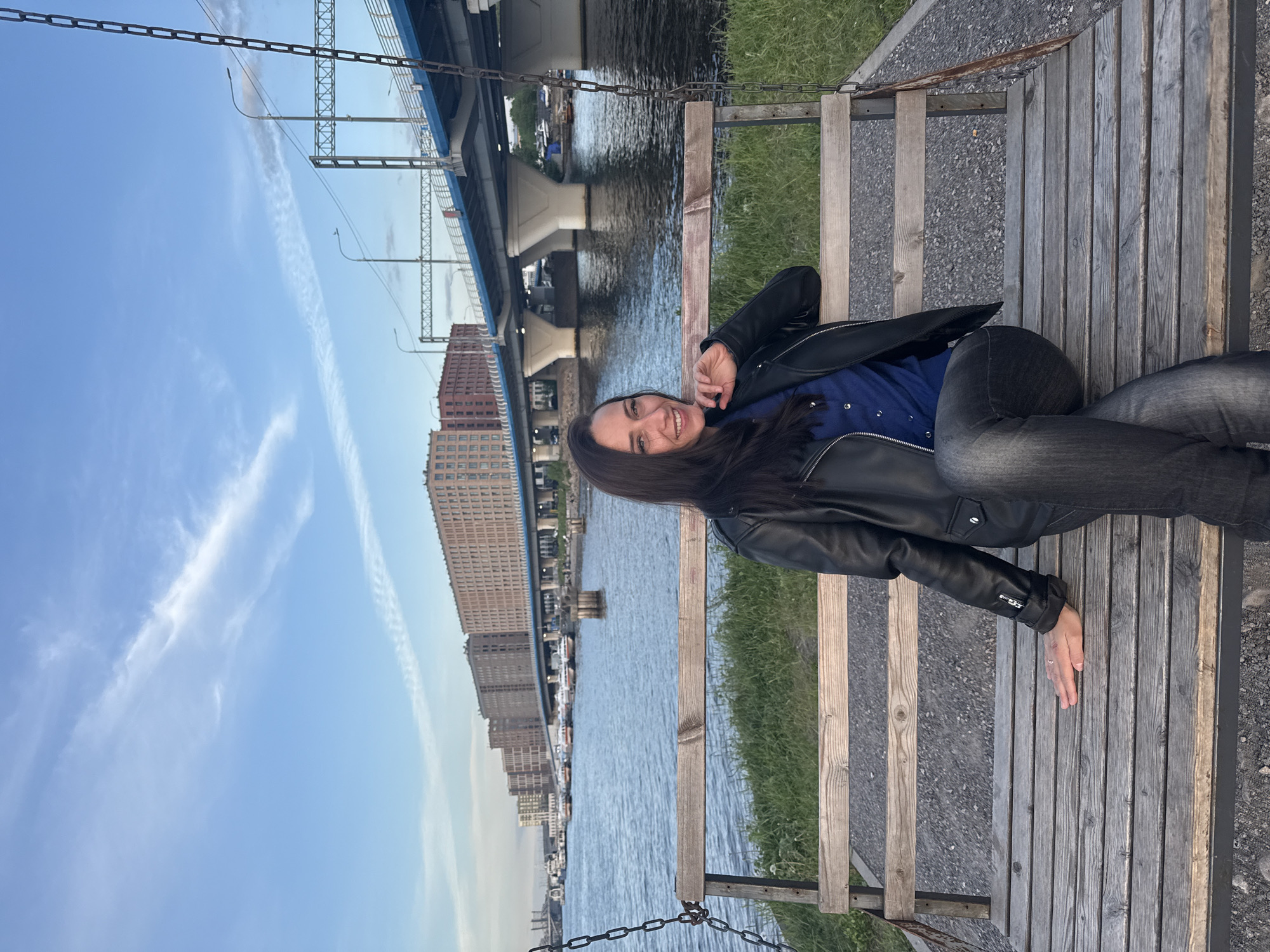 A young woman with long dark hair is depicted in a full-length, casual portrait, lying or reclining on a rustic wooden swing or bench. She is smiling cheerfully at the camera, conveying a sense of relaxation and happiness. She wears a black leather jacket over a blue collared shirt and dark jeans. A simple ring is visible on her left hand. The wooden swing is suspended by metal chains, resting on a gravel path adjacent to a strip of green grass.
In the background, a wide river or canal stretches across the frame, its calm surface reflecting the soft light. On the far bank, several large, multi-story buildings, some with reddish-brown brick exteriors, line the waterfront, with smaller boats visible along the shore. A prominent blue bridge with visible support structures and overhead light posts spans the waterway further back, hinting at a bustling urban environment. The sky above is a clear, light blue with delicate white contrails, suggesting a pleasant late afternoon or early evening. The scene captures a tranquil moment in an urban setting identified as Saint Petersburg, Russia. No visible text is clearly discernible.