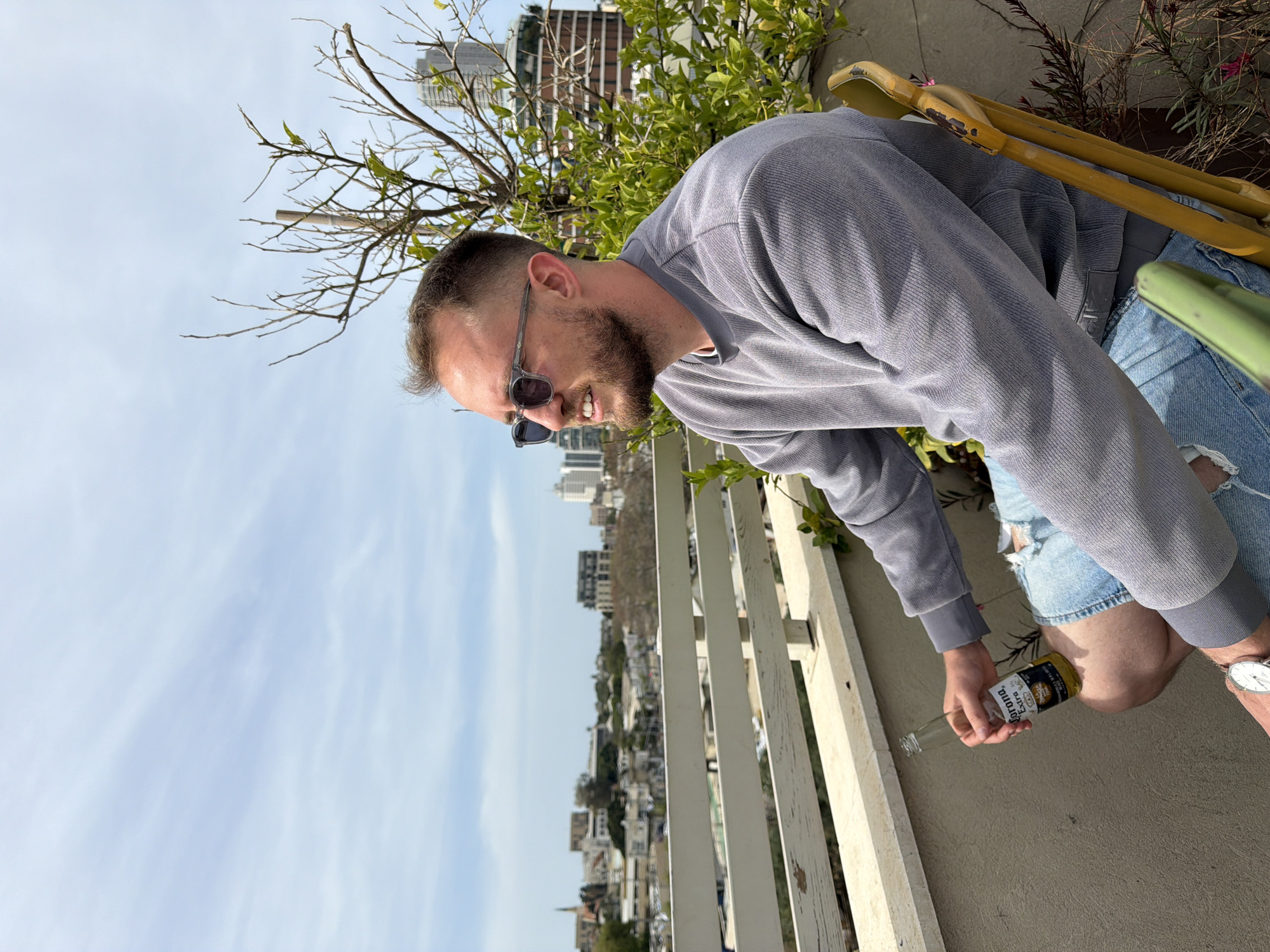 The image captures a man on a sunny day, likely on a balcony or rooftop overlooking a city, identified as Tel Aviv, Israel. The man is seen from the waist up, smiling broadly while looking downwards and slightly to his left. He wears dark sunglasses, a grey long-sleeved top, and light blue ripped denim shorts, with a silver watch on his left wrist.
He holds an empty clear glass bottle of "Corona Extra" beer in his right hand. To the right of the man, a yellow, possibly bamboo, structure is visible, along with green foliage and some dry branches. A white wooden fence or railing extends across the lower portion of the image, separating the foreground from the distant urban landscape.
The background features a clear blue sky with some soft white clouds and a panoramic view of city buildings and residential areas stretching into the horizon, indicative of a vibrant urban setting. The bright light and clear sky suggest it is daytime, possibly midday. The man's smile conveys a relaxed and happy mood.