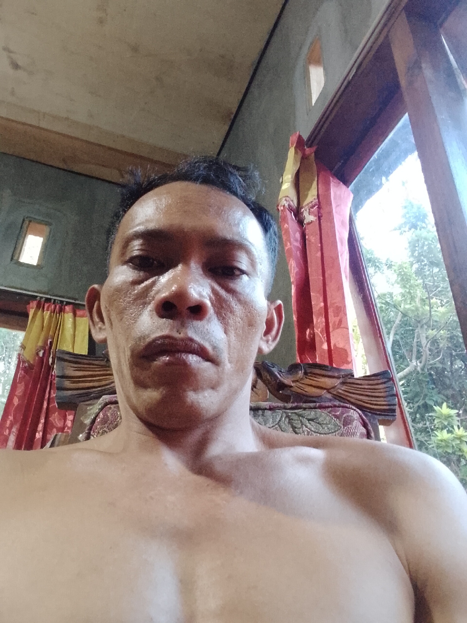 A close-up, low-angle, eye-level, indoor selfie shows a bare-chested man with dark hair, looking directly at the camera. He has prominent cheekbones, a broad nose, and thin lips. His skin appears moist, and there are visible lines around his eyes and mouth. He is seated, with the top of a dark, wooden chair with an intricate design visible behind him. Red and yellow patterned curtains hang to his left, and a tall wooden window frame is on his right, revealing a blurred background of green foliage and bright daylight, suggesting an outdoor environment beyond the room. The room's interior features a ceiling with exposed wooden beams and textured grey walls with small, rectangular windows. The overall lighting is bright, casting shadows on the man's face. The setting is likely Madukoro, Indonesia, given the context.