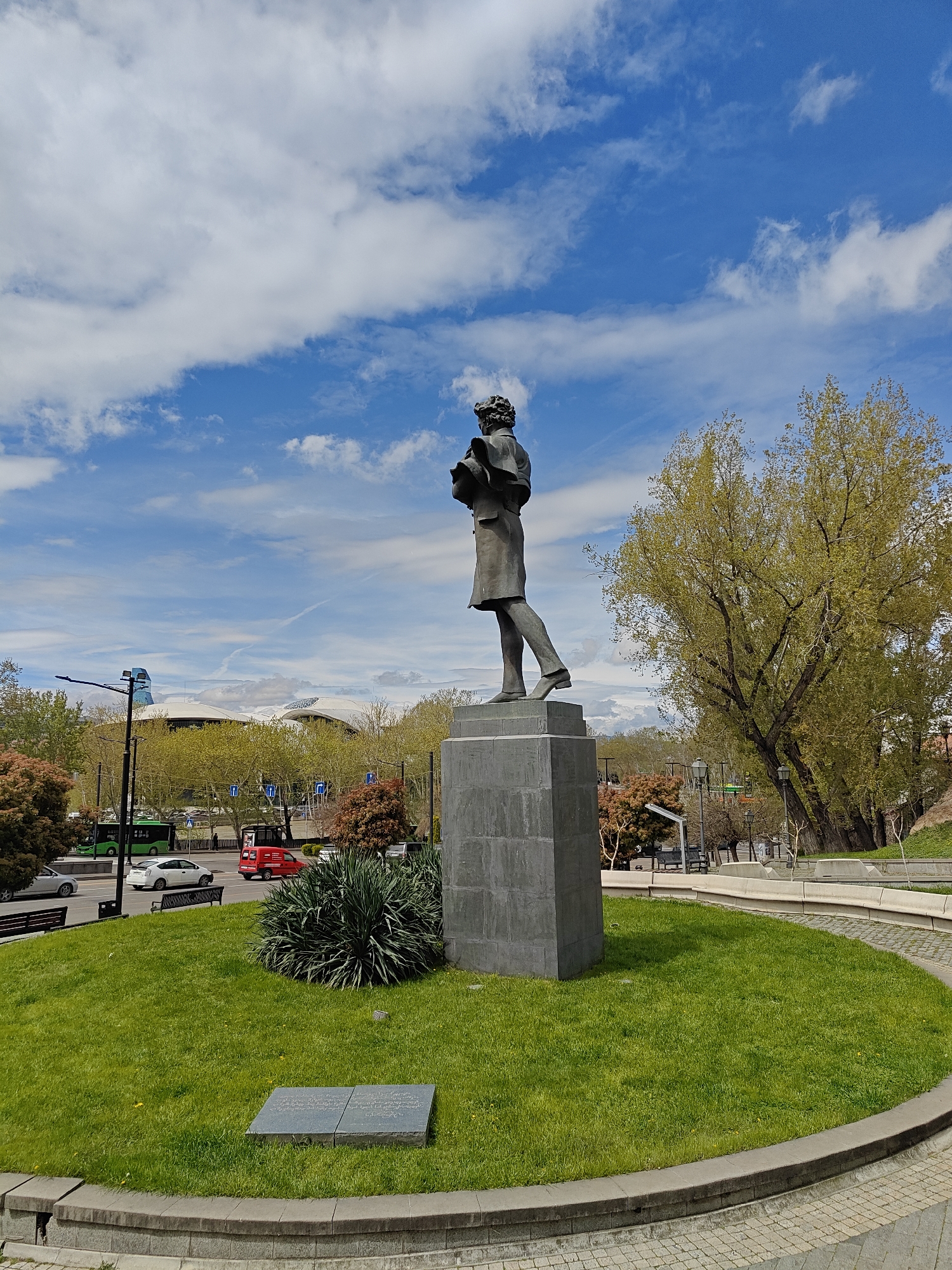 The image depicts a bronze statue of a man standing on a grey stone pedestal, positioned on a circular patch of green grass within a landscaped park in Tbilisi, Georgia. A rectangular grey plaque lies flat on the grass in front of the statue. The statue portrays a man in period clothing, with a jacket and trousers, holding an object in his hands, possibly a book.
In the background, a multi-lane road is visible with various vehicles including a green bus, white car, and red van. Beyond the road, trees line the street. In the distance is a modern architectural structure with a curved, white roof. The sky is blue with scattered white clouds. A large tree with green leaves stands to the right of the statue. The park is edged with stone walls and a paved path.