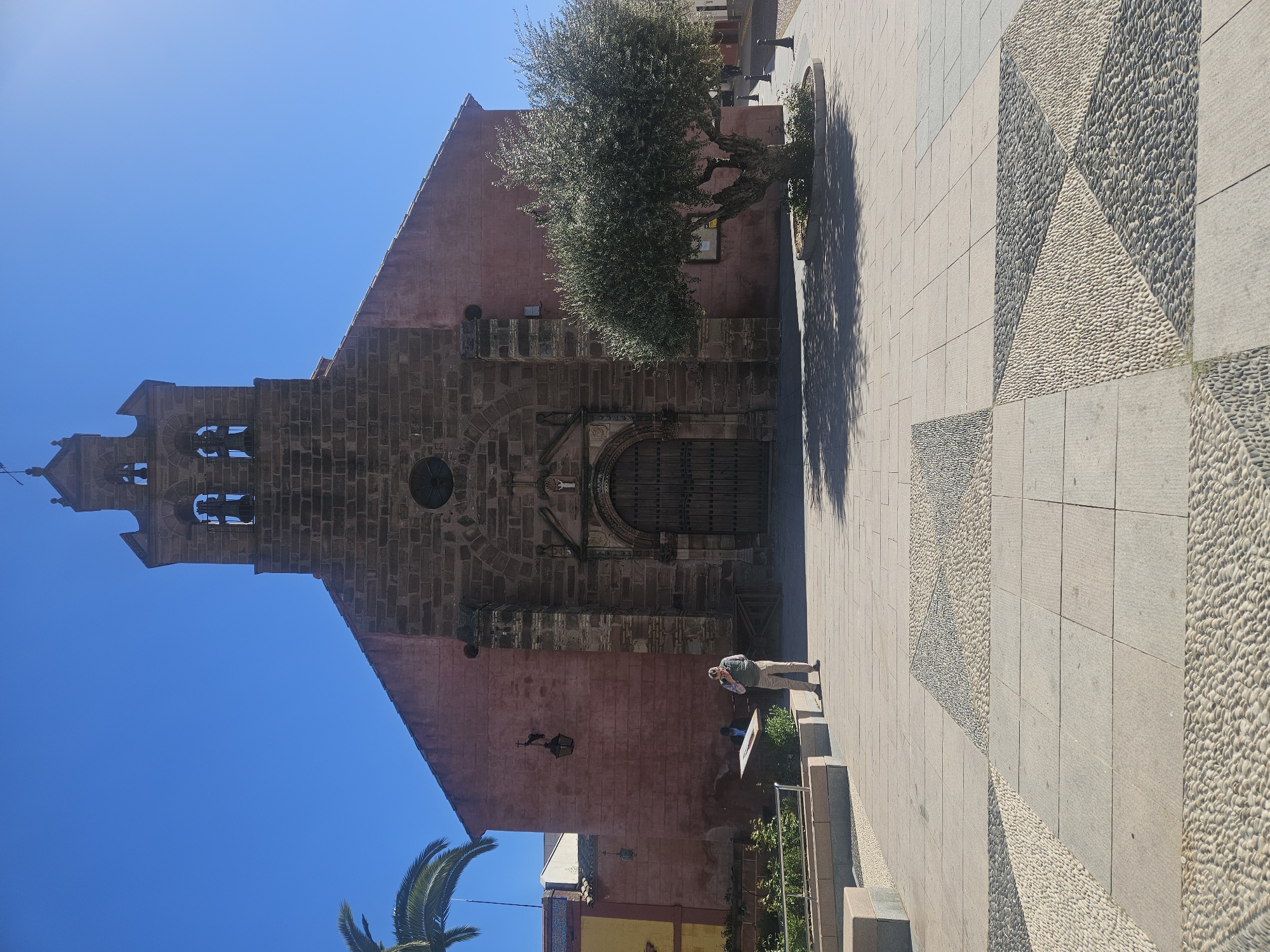 Here is a description of the image:
The image is a medium shot of a church in Pedro Abad, Spain. The church is made of red brick and has a tall bell tower. A tree is to the right of the church, obscuring some of the façade. A person is standing in front of the church, looking at a sign. The ground is paved with stone, and the sky is blue. The paving pattern has a geometric pattern of light stones on darker stones.