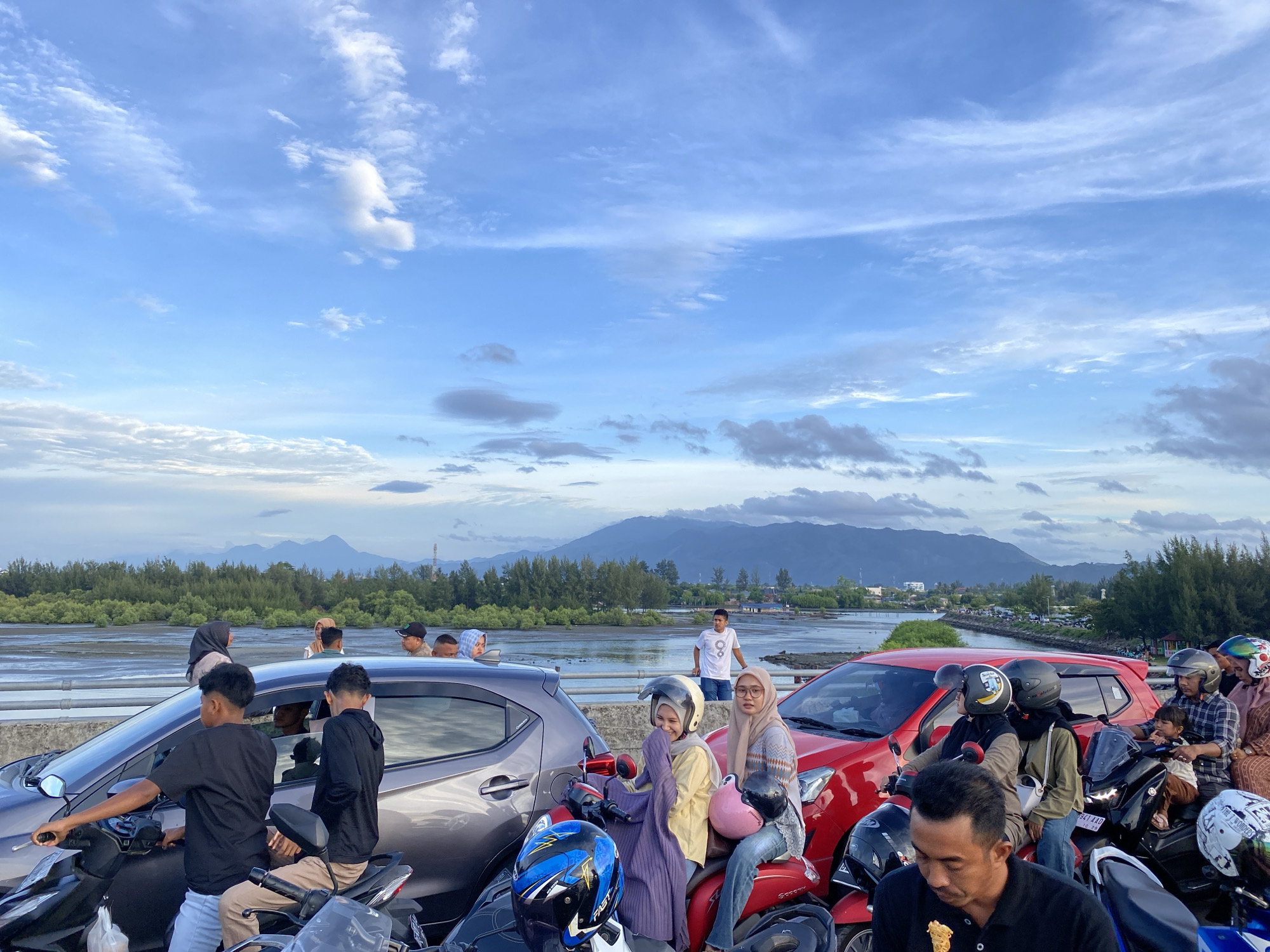The image captures a scene at the Ulee Lheue Bridge in Banda Aceh, Indonesia. A diverse group of people are gathered, many of them riding motorcycles or sitting in cars, suggesting a communal gathering or a popular viewpoint. The sky above is filled with dynamic clouds, indicative of a potentially changing weather pattern, with a bright blue expanse peeking through.
In the foreground, several motorcycles are parked, some occupied by individuals. One motorcycle in the lower left has a rider in a black t-shirt and white pants, seemingly preparing to move. Next to him, another rider in a black hoodie is seated. Further to the right, a red car is prominently featured, with several individuals around and on motorcycles in front of it. Two women are seated on a motorcycle in front of the red car, one wearing a yellow outfit and the other a beige headscarf and patterned sweater.
The background reveals a picturesque landscape with a body of water, possibly a river or the sea, bordered by lush green trees that line the shore. Beyond the trees, a mountain range stretches across the horizon, partially obscured by atmospheric haze. The overall impression is one of a lively, community-oriented setting within a natural and scenic environment.