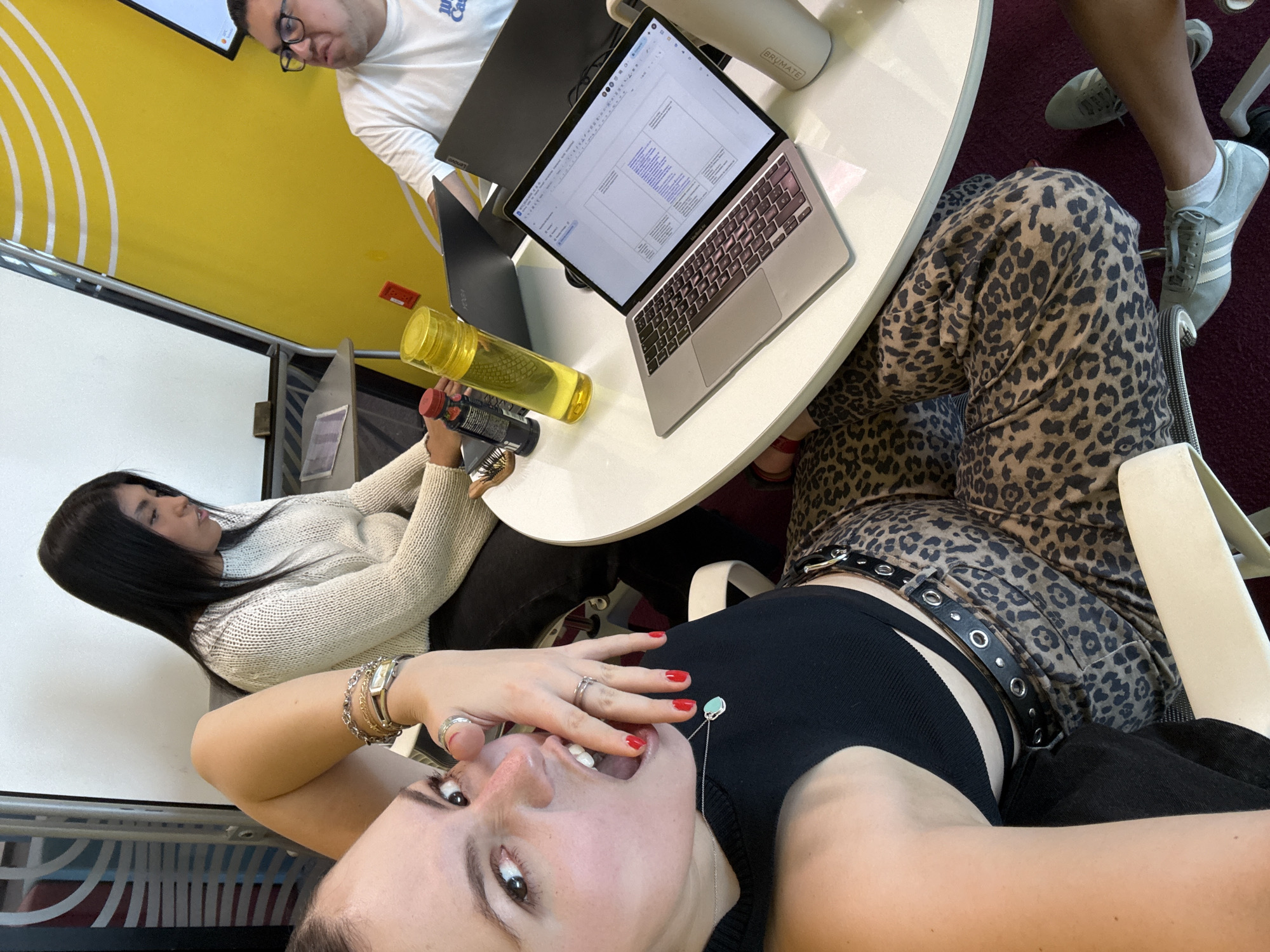 An indoor, high-angle selfie captures three individuals engaged in what appears to be a collaborative workspace in Mexico City, Mexico. The perspective is from the bottom right, looking up and across the scene.
In the immediate foreground, a person with dark hair and fair skin is taking the selfie. They are wearing a black ribbed tank top, leopard print pants, and a black belt with silver grommets. Their hand, adorned with multiple rings and red nail polish, is near their open mouth, which has the tongue slightly out, conveying a playful or expressive emotion. A silver necklace with a light blue pendant hangs around their neck.
To the left, another person with long dark hair wears a cream-colored knit sweater. They are seated at a white round table, looking down at their phone while holding a black bottle. A clear yellow water bottle is also on the table.
Further back, a third person, wearing glasses and a white T-shirt with the partial text "Just Cause" or similar, is seen leaning over a dark-colored laptop, possibly a Lenovo. On the white table, an open silver MacBook Air displays a document with structured text or a presentation. A white bottle with the visible brand name "BRUMATE" is also present.
The background features a vibrant yellow wall with white curved stripe patterns, and a white board is visible on the left. The floor is covered in a red carpet. The attire of the individuals is casual, suggesting a relaxed yet focused work or study environment. The time of day is not explicitly indicated but appears to be daytime.