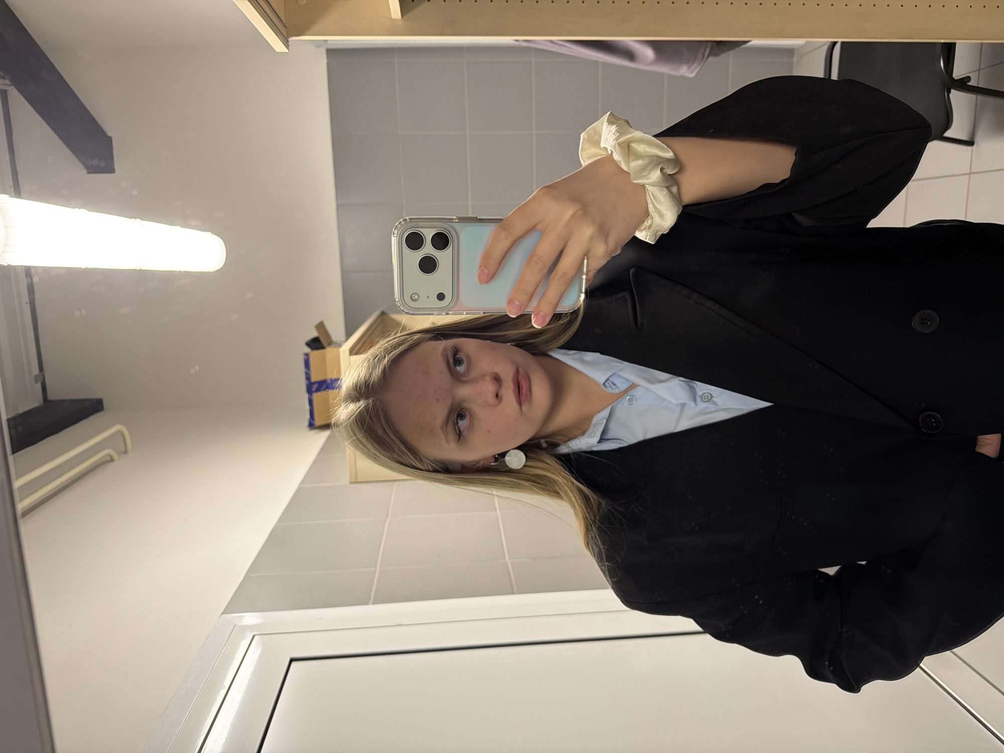 A young woman is taking a selfie in a bathroom. She is holding an iPhone with a clear case in her left hand. She is wearing a light blue collared shirt under a black blazer. A cream-colored scrunchie is on her wrist. Her hair is blonde and parted on the side. She is wearing white earrings. The bathroom has white walls and tiled walls. A fluorescent light fixture is visible on the ceiling. A shelf is visible above the tiled wall. A mirror is on the left side of the frame. The location appears to be Izhevsk, Russia, based on common architectural and interior design elements.