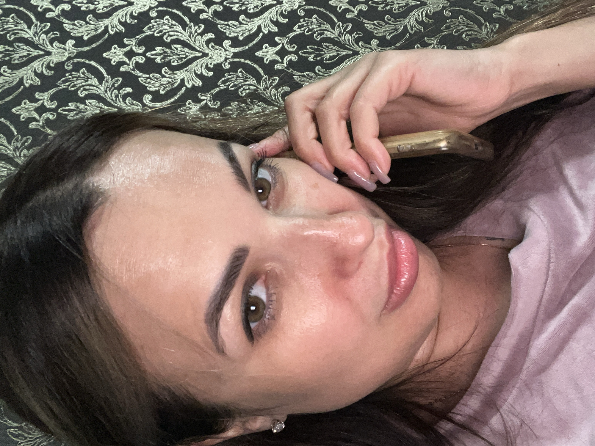 A medium close-up selfie shows a woman holding a gold-colored smartphone to her right ear. She is looking directly at the camera with a slightly pensive expression. Her dark brown hair is pulled back loosely, with some strands falling around her face. She is wearing a light pink, velour top and a thin gold necklace. A small diamond stud earring is visible in her left ear. The background features a dark wallpaper with an ornate, light gold damask pattern. The lighting appears to be artificial, casting a soft glow on her face and accentuating the sheen on her skin.