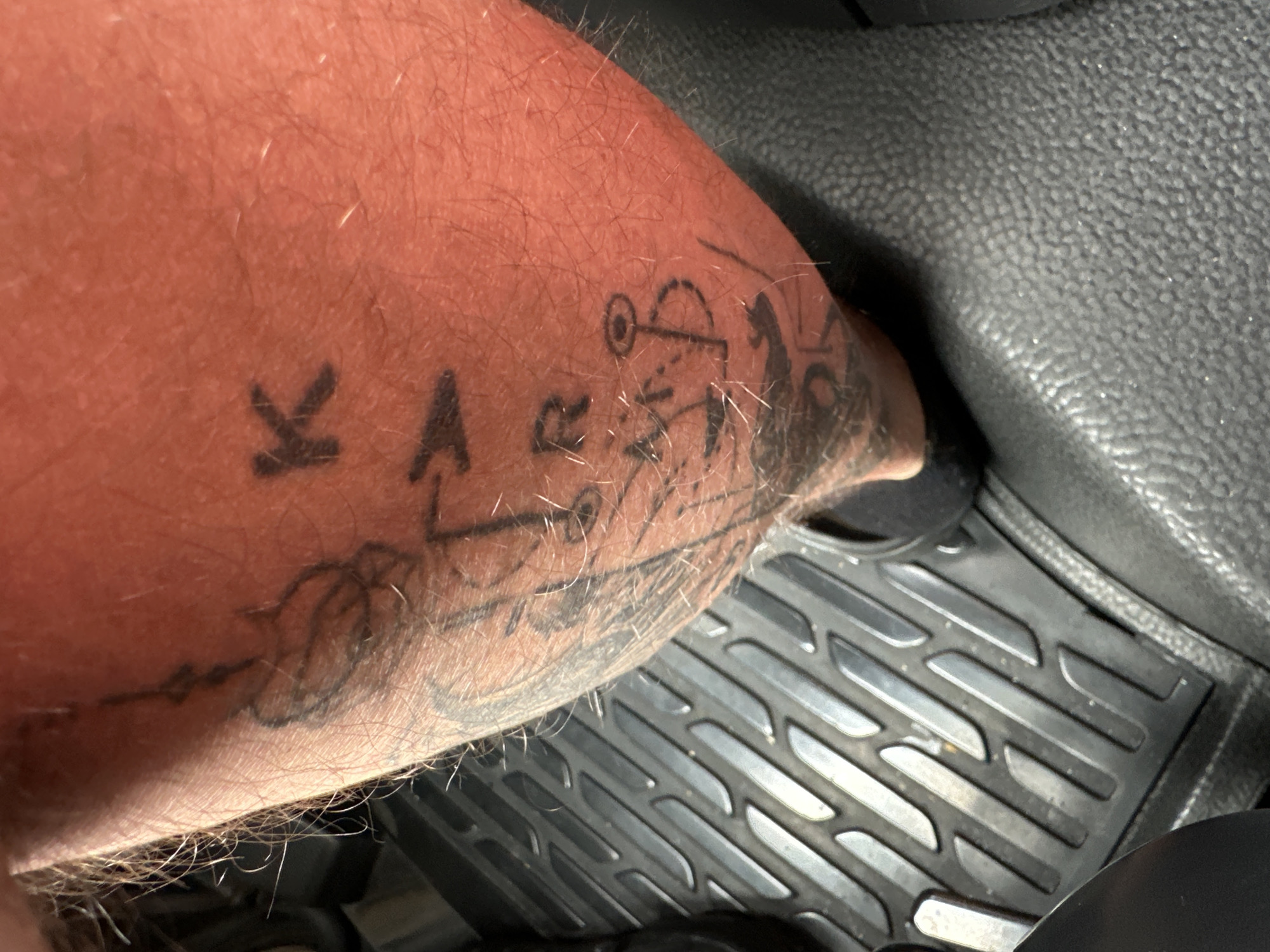 The image shows a close-up view of a person's calf with a tattoo. The tattoo appears to be of the word "KARAA" written vertically in black ink, with some additional symbols or drawings beneath it that are less distinct. The skin is fair and has some visible hairs. The person is wearing a dark-colored shoe. The background shows a textured, dark grey rubber floor mat, likely from a vehicle. The scene is set in an indoor environment, potentially within a car or a similar space with floor mats. There is no indication of the city of Nossen, Germany, in the image itself.