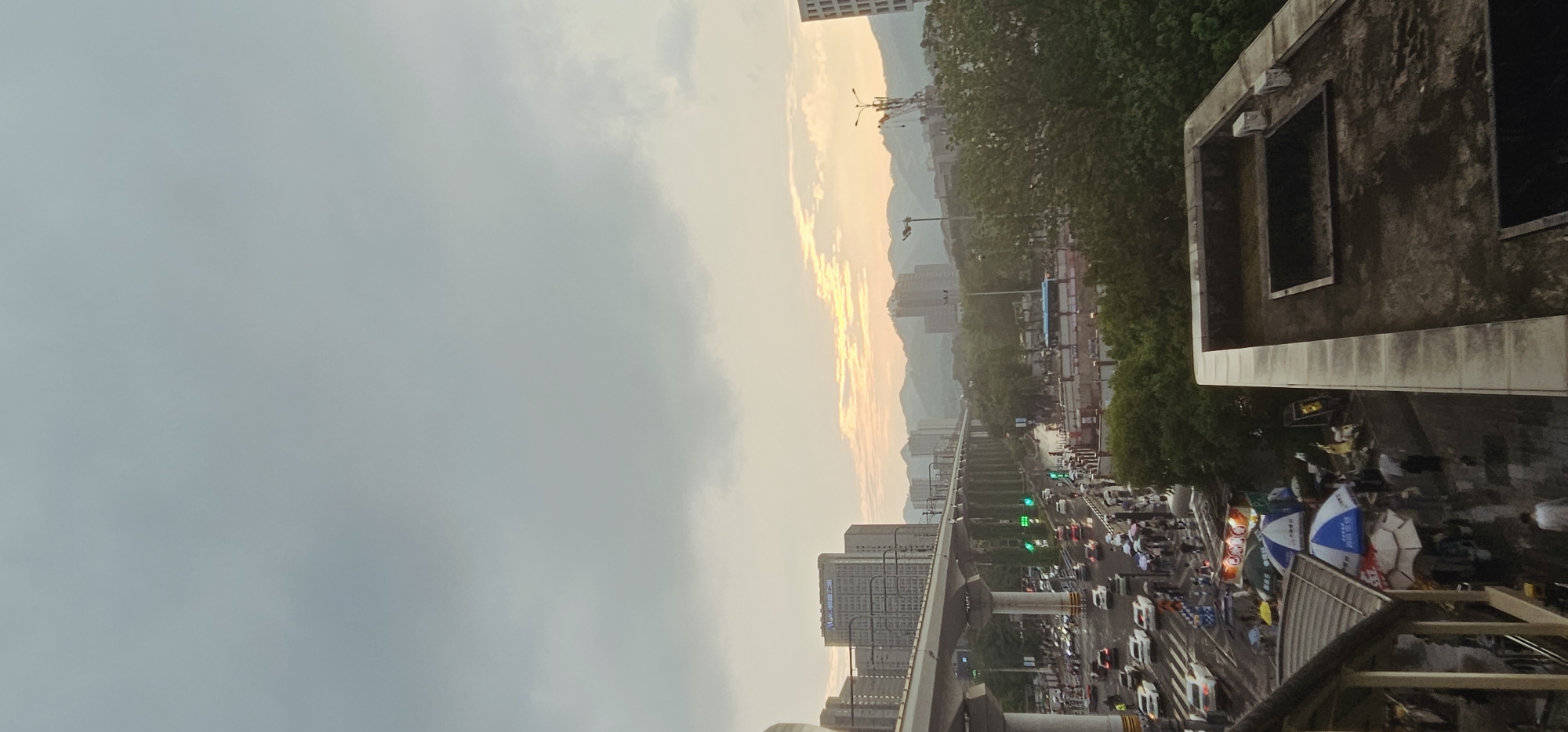 The image depicts an evening scene in Chongqing High-tech Zone, China, showing a busy urban environment. A partly cloudy sky with an orange glow dominates the upper portion of the frame. Mountains and buildings are visible in the distance. A monorail track runs alongside a street filled with traffic. Multiple cars, and pedestrians with umbrellas are visible on the street level. A concrete building structure with visible window openings is in the foreground on the right.