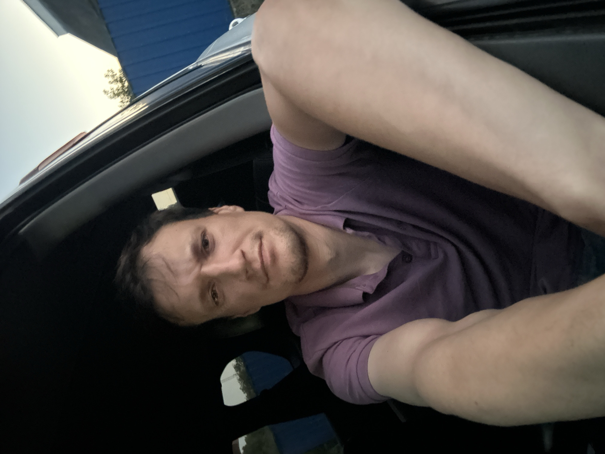 A man sits in the driver's seat of a car. He is wearing a purple polo shirt and has short brown hair. His arms are resting on the steering wheel, and he is looking at the camera. The car is parked outdoors, with a blue wall and some trees visible in the background. The sky is partly cloudy.