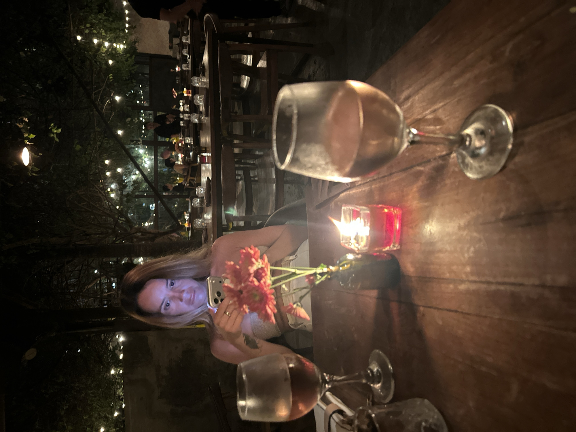 A medium shot photograph shows a woman with blonde hair sitting at a wooden table in an outdoor dining area, illuminated by string lights and a candle. She is holding a smartphone in her hands and looking at it. In front of her is a glass of rosé wine and a small vase with pink flowers. Another glass of rosé wine is in the foreground. The background shows several other tables set with glassware and candles, and people dining. The setting appears to be a restaurant in Kuta Selatan, Indonesia.