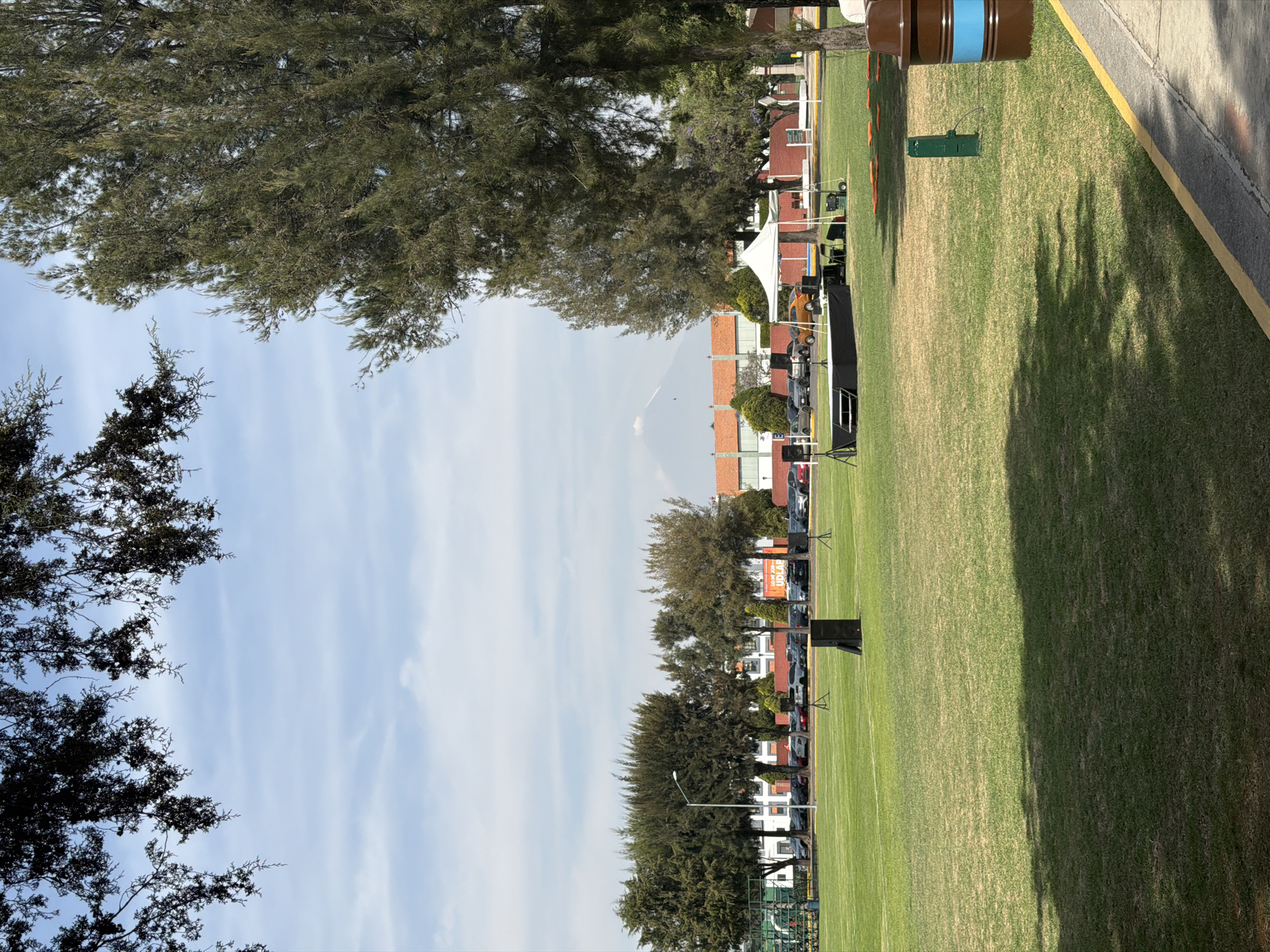The image depicts a green field with trees lining the left side and buildings in the background. In the middle of the field, there is a stage set up with a white tent, speakers, and other equipment. Vehicles are parked alongside the stage. Cones are seen placed near the top of the field and it's adjacent to a building with a yellow border. In the distance, a mountain is faintly visible. The location is in the city of San Andrés Cholula, Mexico.
