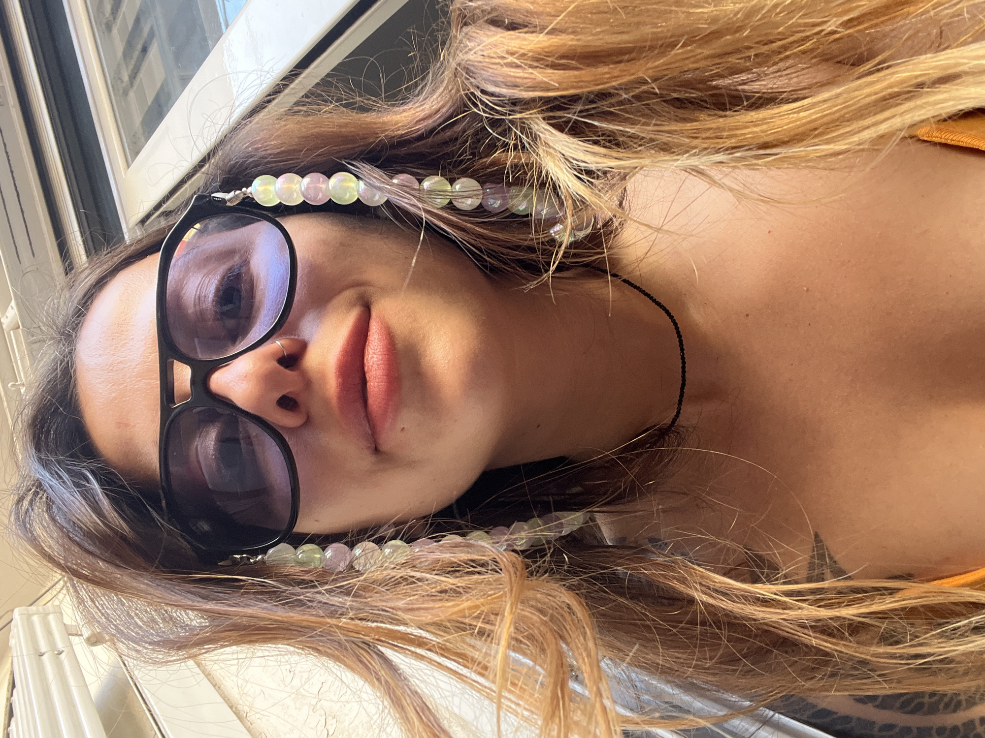 The image is a close-up, horizontally oriented portrait of a person, likely a woman, with her head positioned towards the left of the frame. Her face is angled slightly upwards, with her gaze directed towards the viewer through dark sunglasses. Her long, light brown and blonde hair, styled with loose waves, spreads out around her head and across her shoulder area. She wears black-framed sunglasses that reflect a bright, indistinct light source. A decorative glasses chain, featuring iridescent round beads in shades of pink, green, and clear, is draped around her head, intermingled with her hair. A thin black cord is visible around her neck. Her lips are slightly parted, revealing a natural pink hue. Part of her shoulder and upper chest are visible on the right, suggesting she is wearing an orange-colored top, and a dark, possibly tattooed, pattern is faintly discernible on her lower shoulder/upper arm. The background is brightly lit, showing blurred window frames and what appears to be an urban building exterior, indicating an indoor setting near a window. The strong natural light suggests it is daytime. The overall expression is relaxed and casual. The location, according to the provided context, is Barcelona, Spain. There is no visible text in the image.
